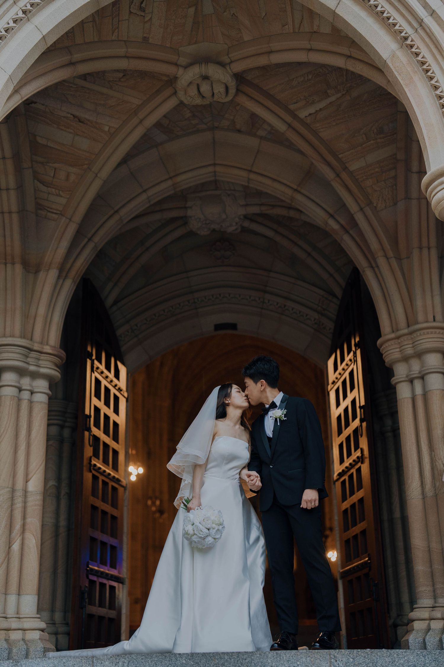 Newlyweds share a romantic moment under ornate stone archways in a historic church corridor.