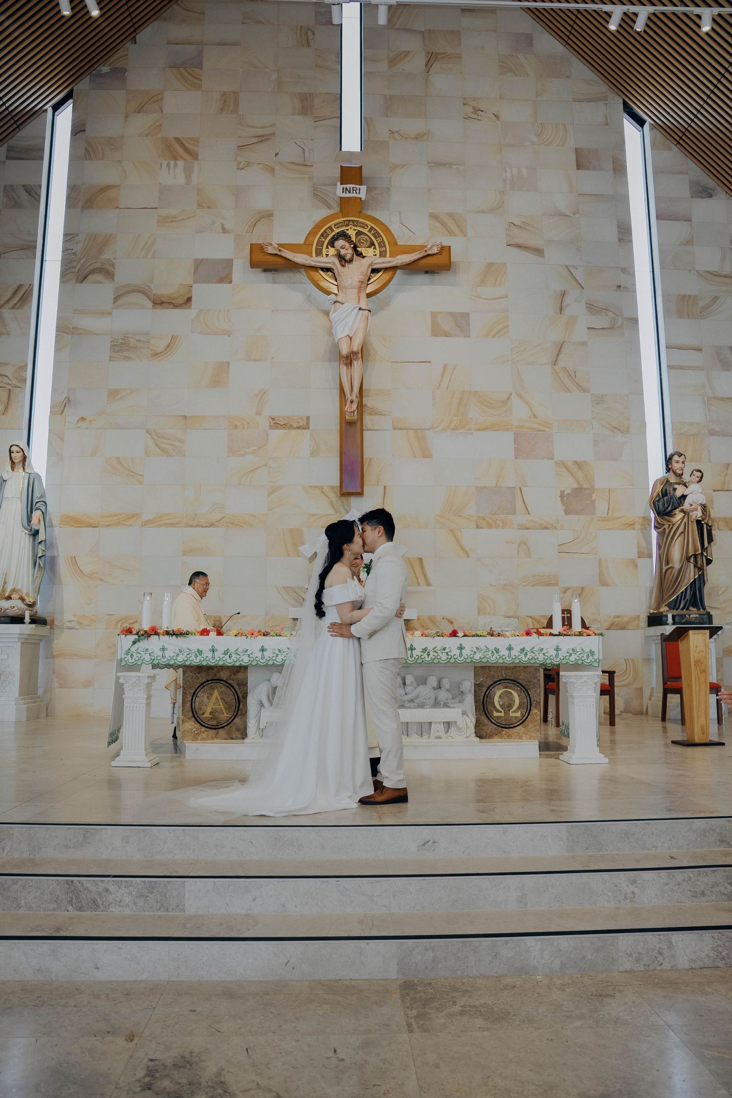 A couple embraces at the altar of a modern church with a large crucifix mounted on the stone wall behind them.