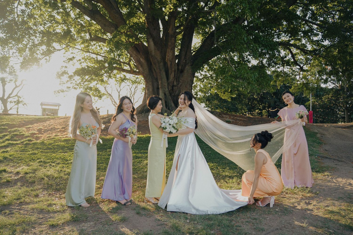 A bridal party in pastel dresses gathers under a large tree during golden hour as they help adjust the wedding veil.