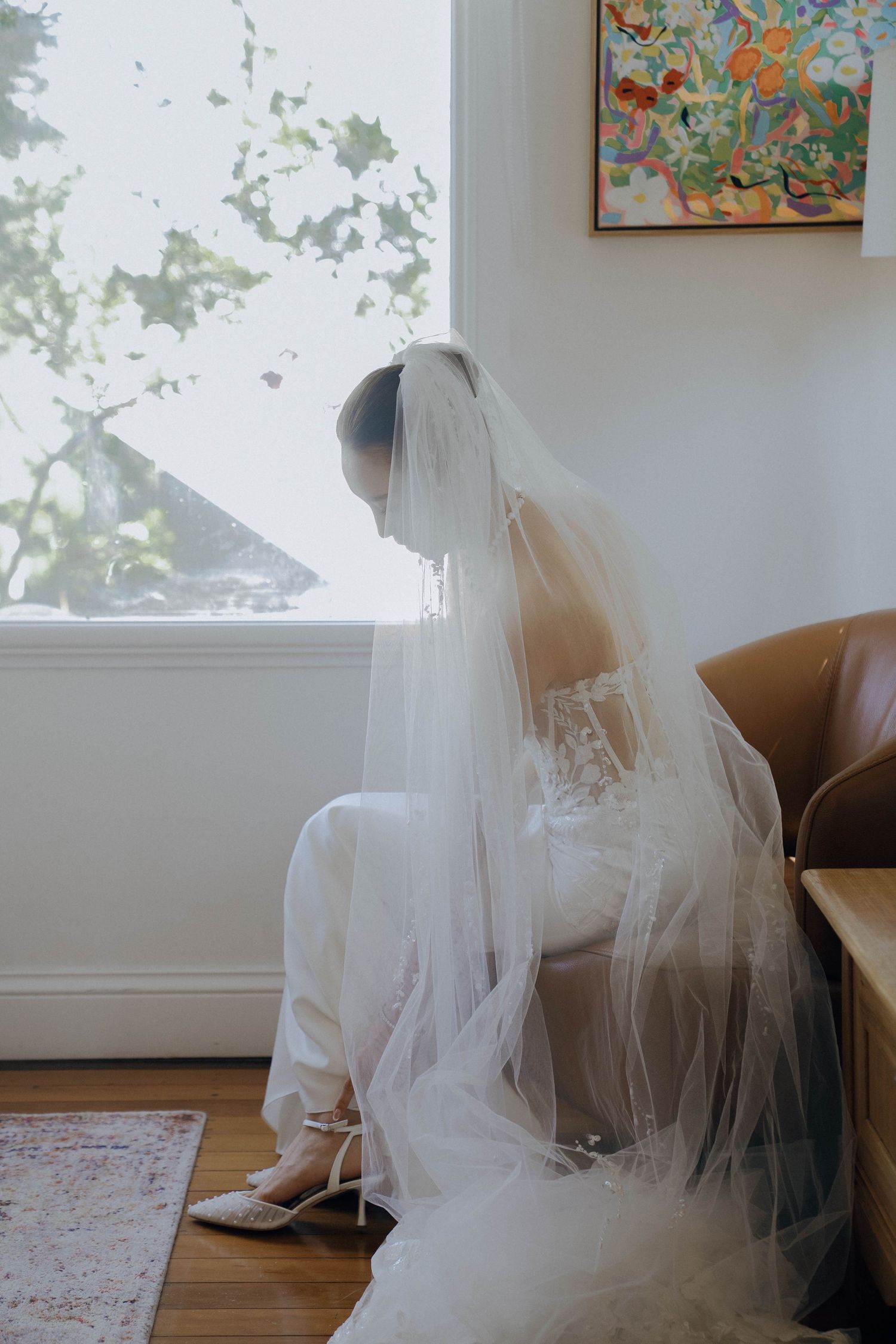 A bride in a flowing white veil and gown sits by a window with natural light streaming in, creating a serene moment.