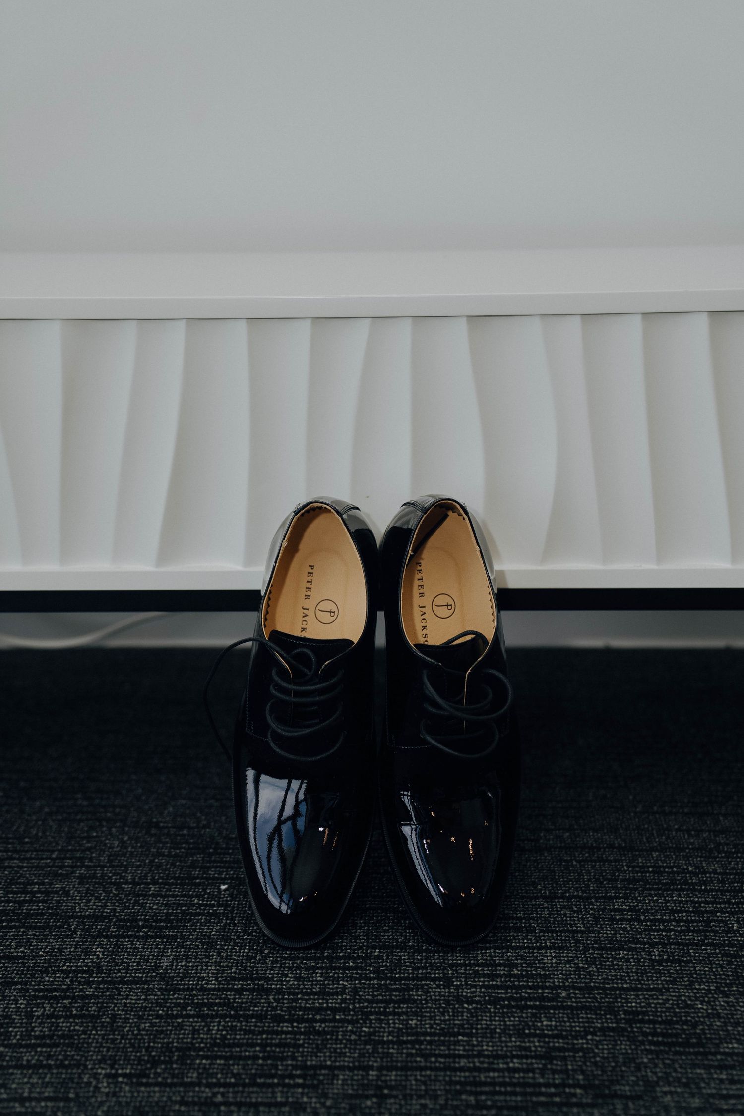 Black leather dress shoes arranged in a row against a dark carpet and white baseboard.