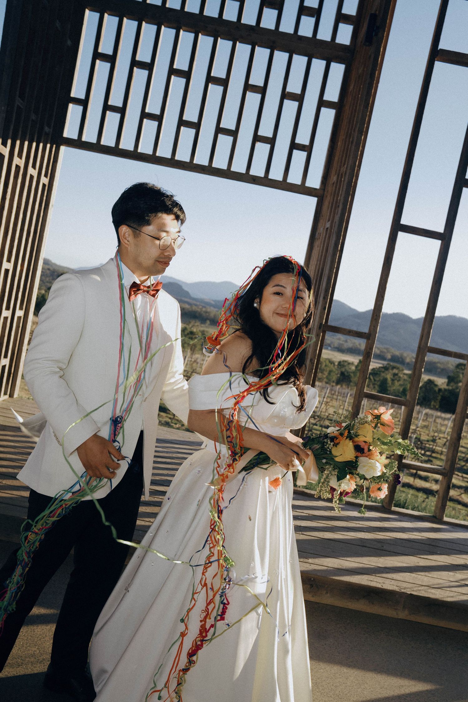 A couple in wedding attire dances together on a terrace overlooking scenic mountain views on a sunny day.