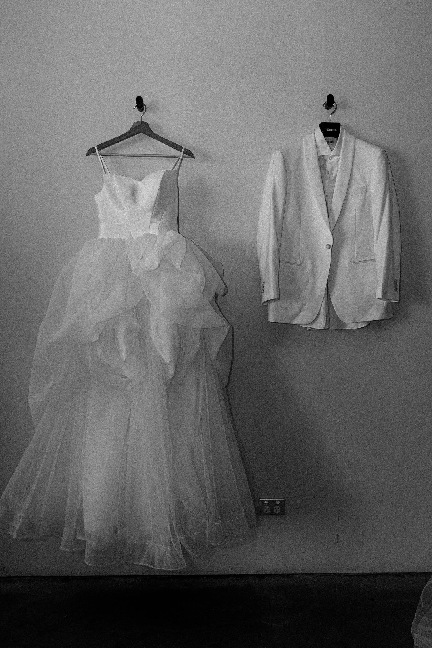 Black and white photograph of wedding attire including a flowing tulle dress and formal suit jacket hanging on wall.