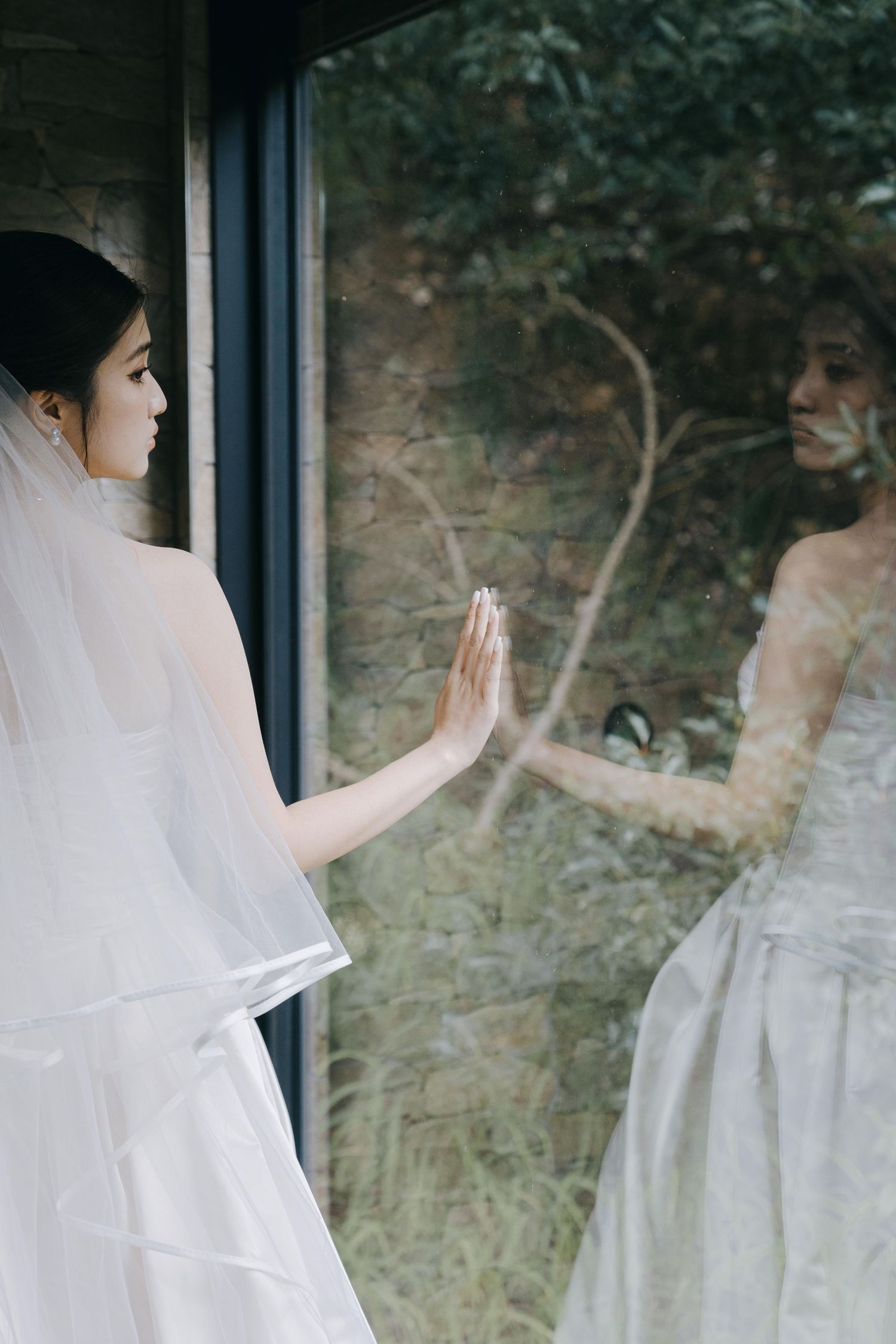 A bride in a white wedding dress and veil touches her reflection in a window with trees visible outside.