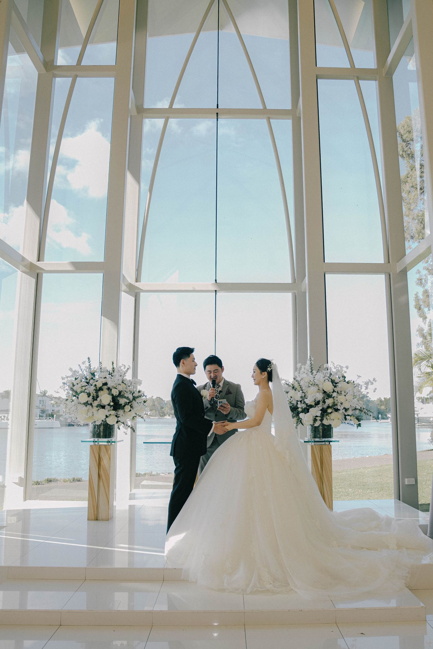 A romantic wedding ceremony in a modern glass chapel with white floral arrangements and water views in the background.