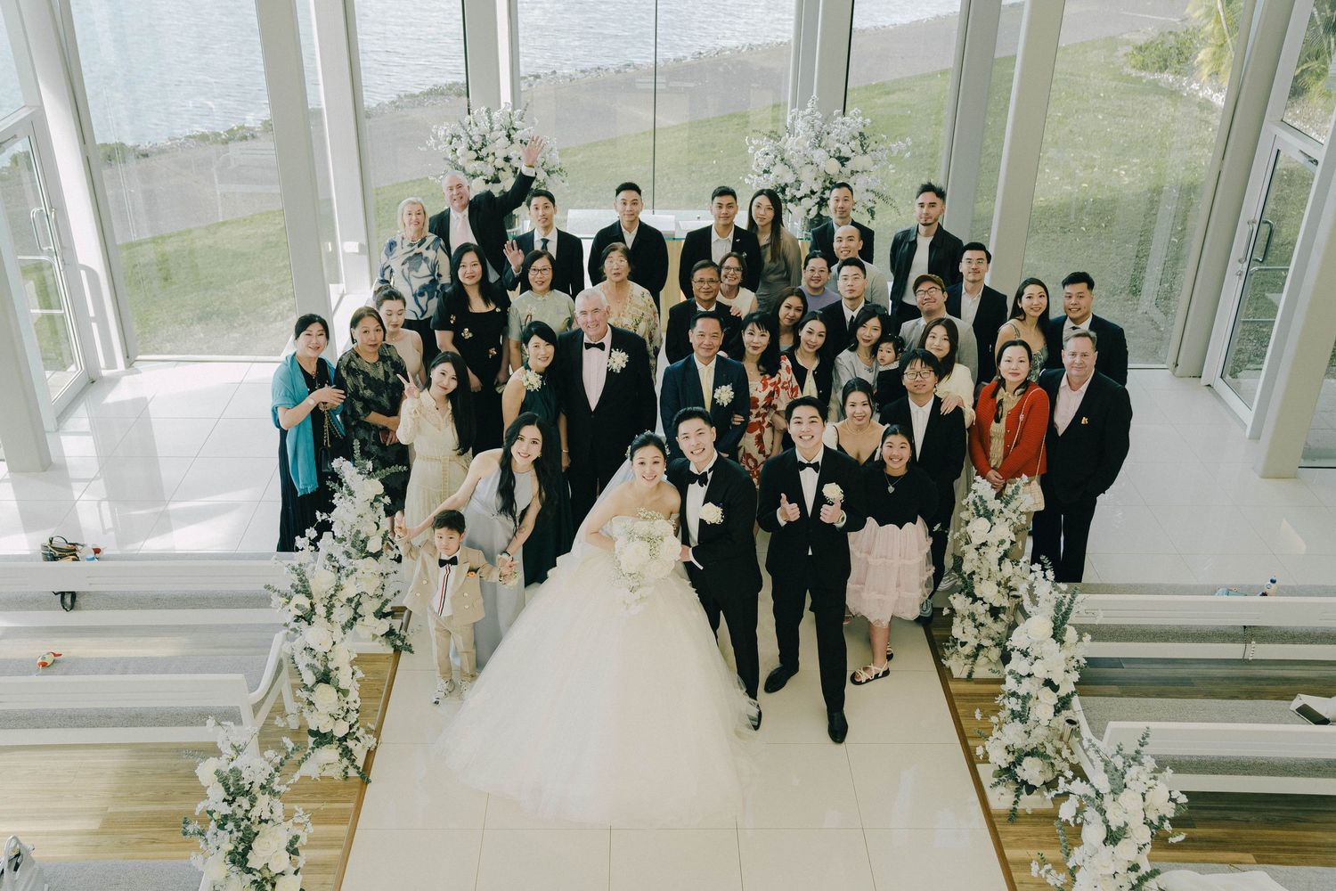 Wedding party gathers in an elegant glass-walled venue with white floral decorations for a formal celebration.
