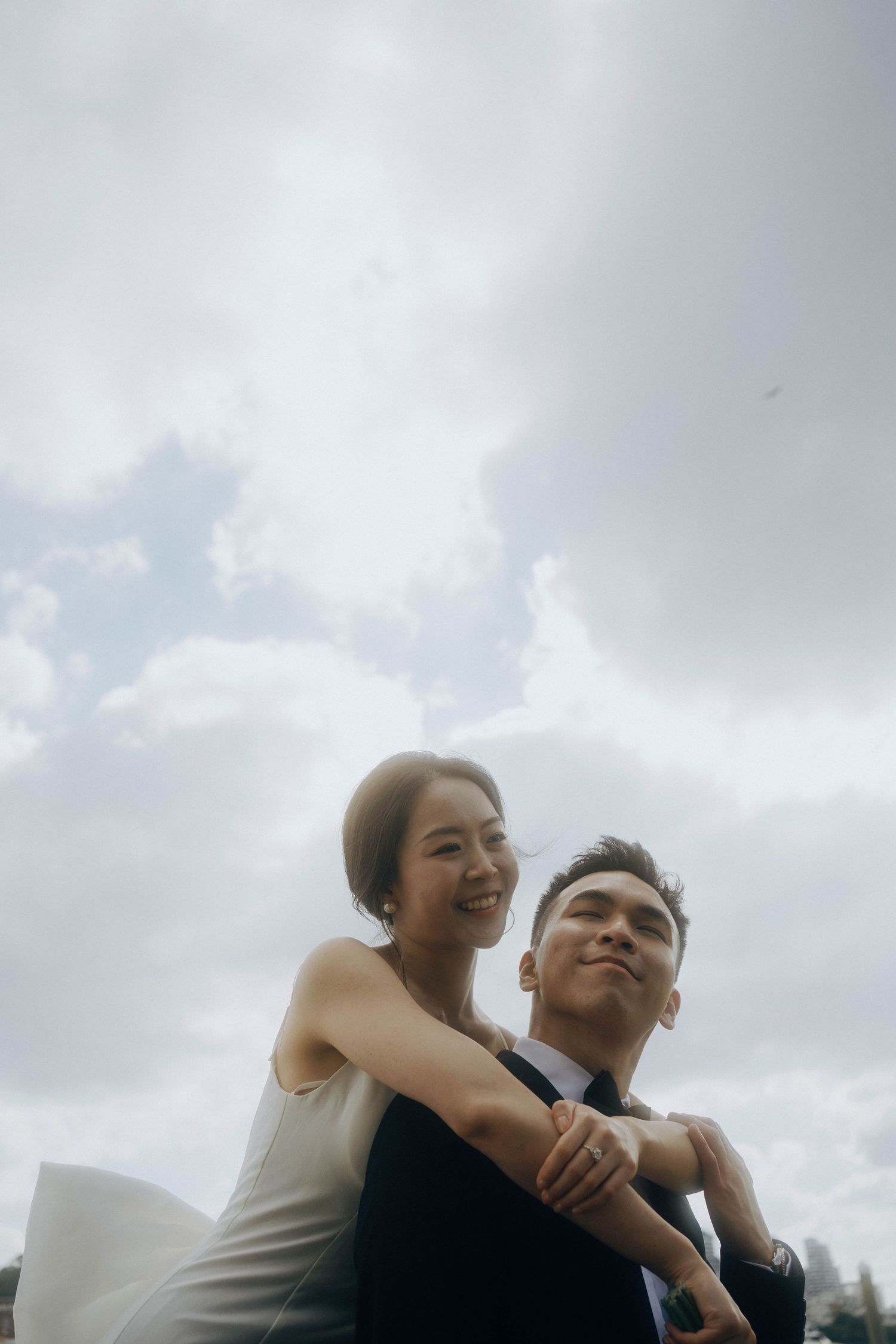 A couple embraces and laughs together under cloudy skies in a candid romantic moment.