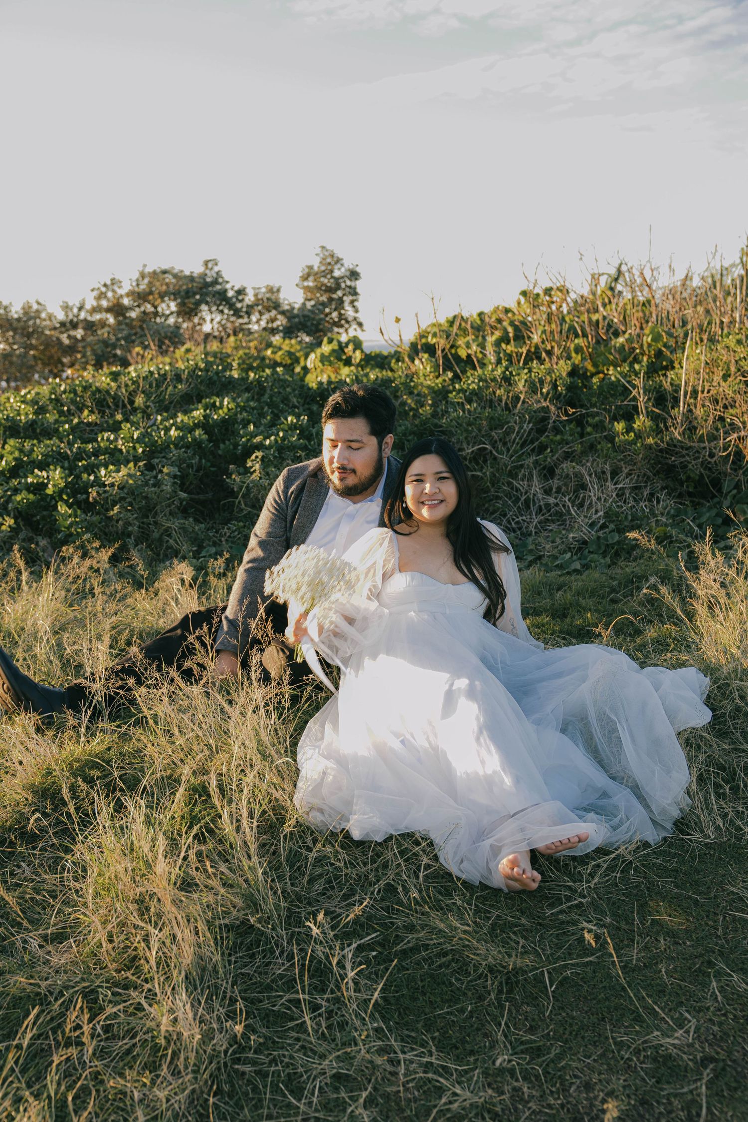 A couple in wedding attire lies together in a grassy field during golden hour.