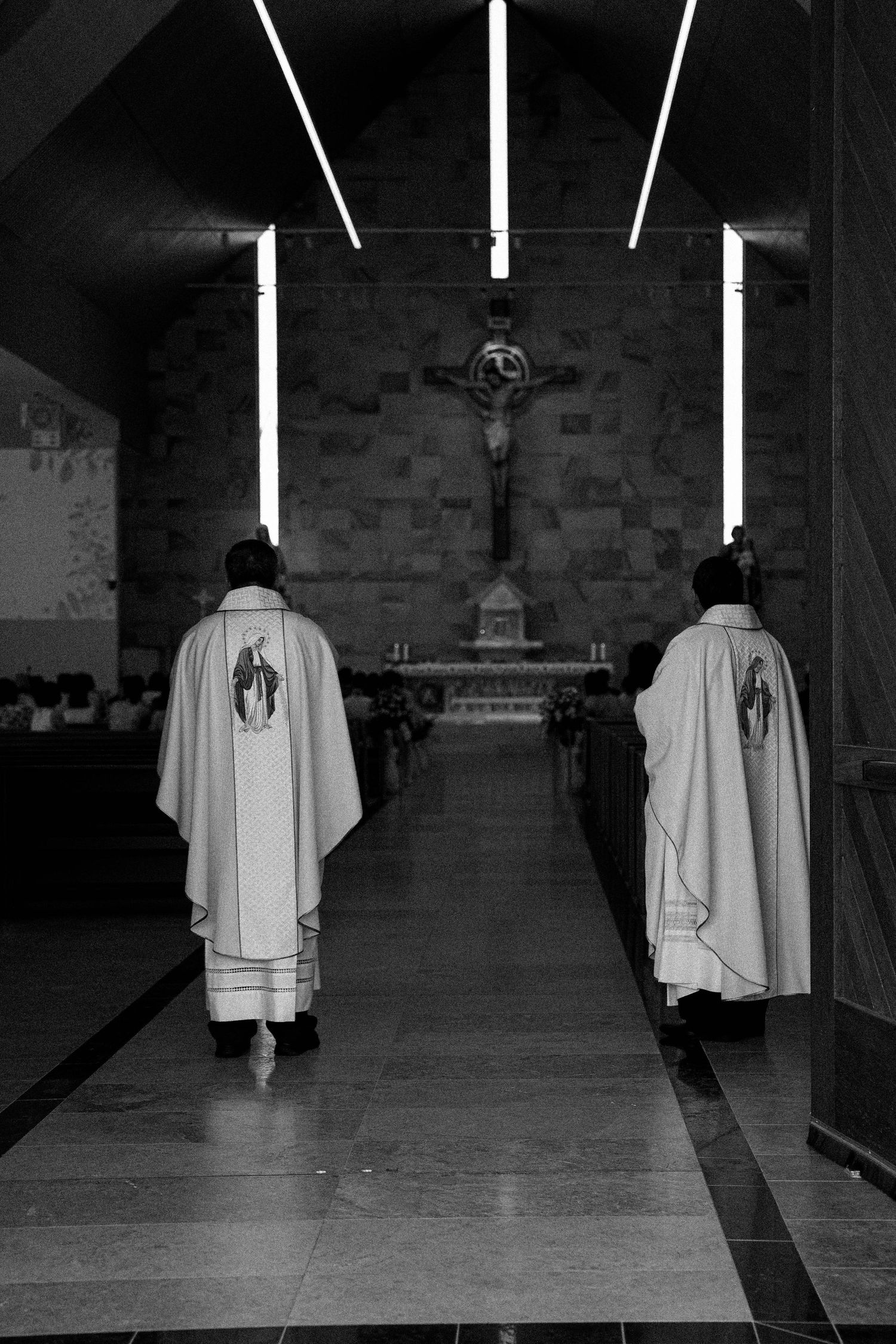 Two priests in white robes walk down church aisle towards crucifix altar in black and white photograph.