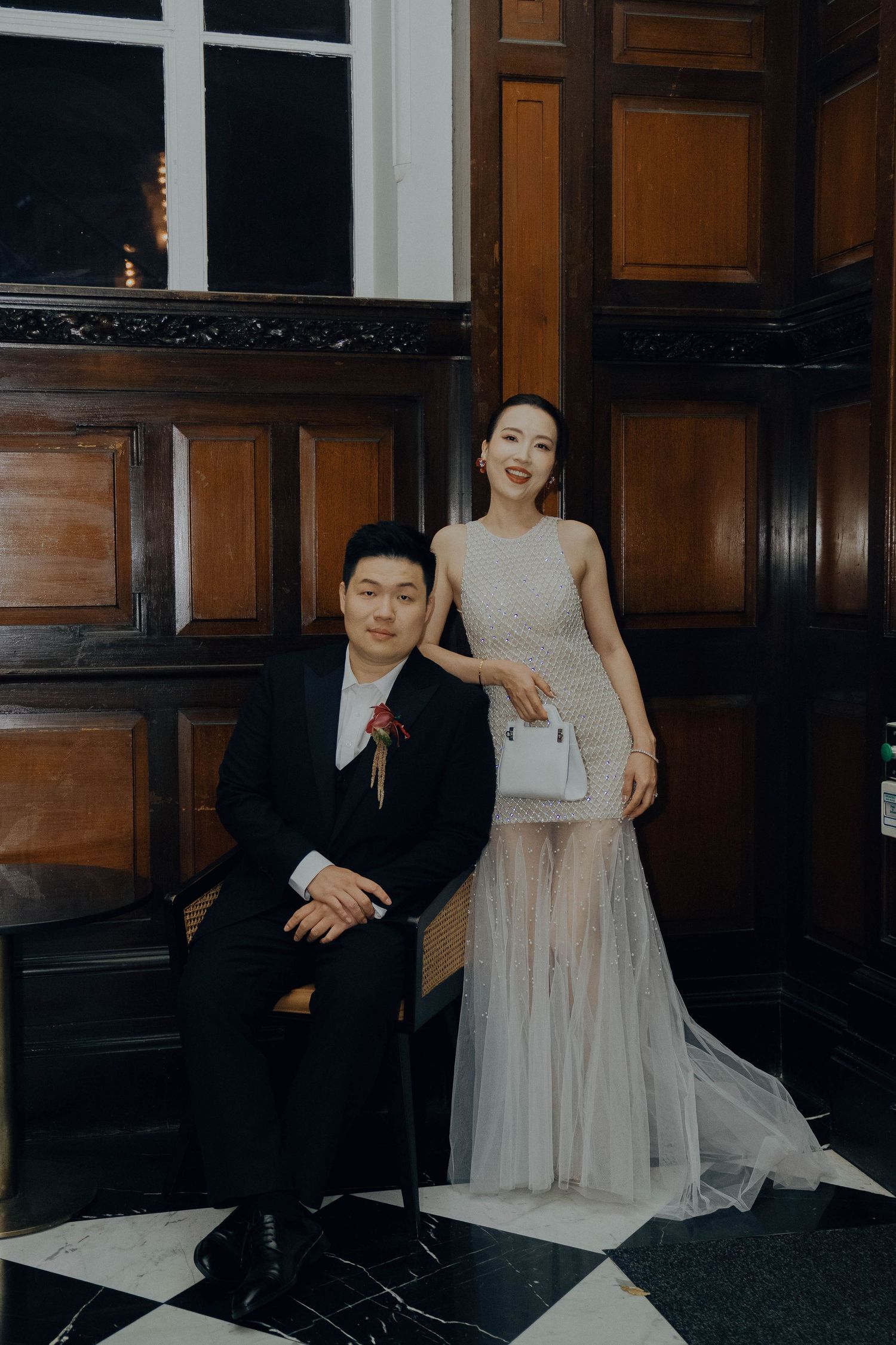 A couple in formal attire poses together in an elegant dark wood-paneled room.