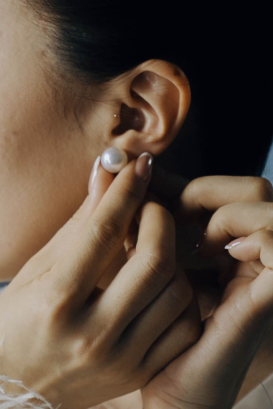 Close-up view of hands adjusting a pearl earring on an ear against a dark background.
