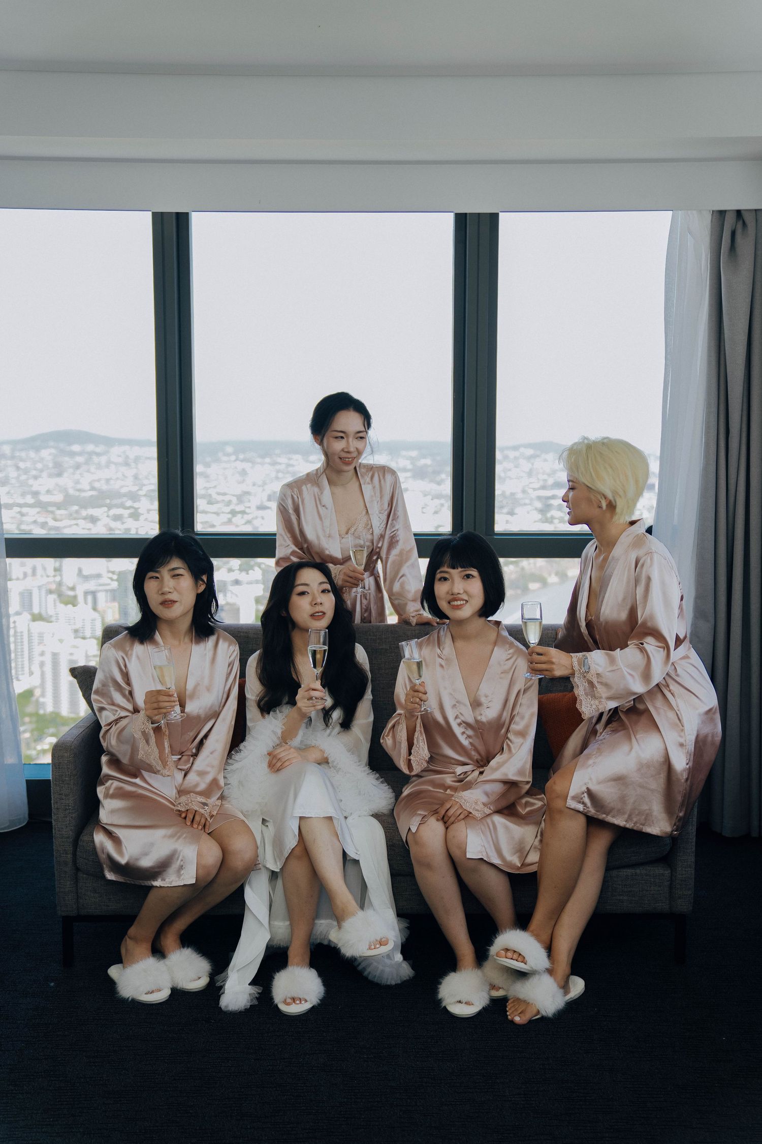 A group of friends wearing matching pink satin robes sit together on a couch with a city view in the background.