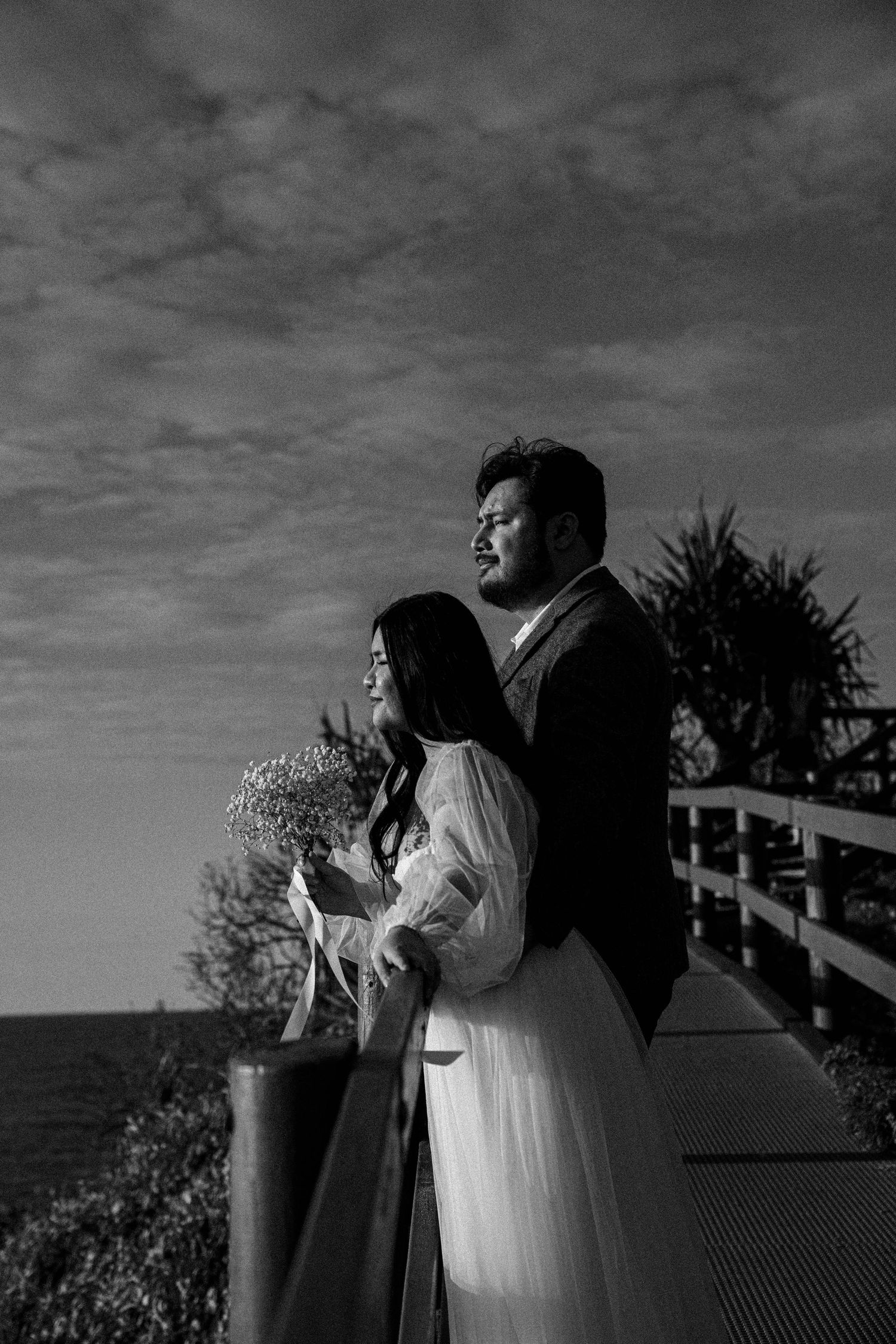 A romantic black and white photo of a couple standing together on a balcony overlooking the ocean at sunset.