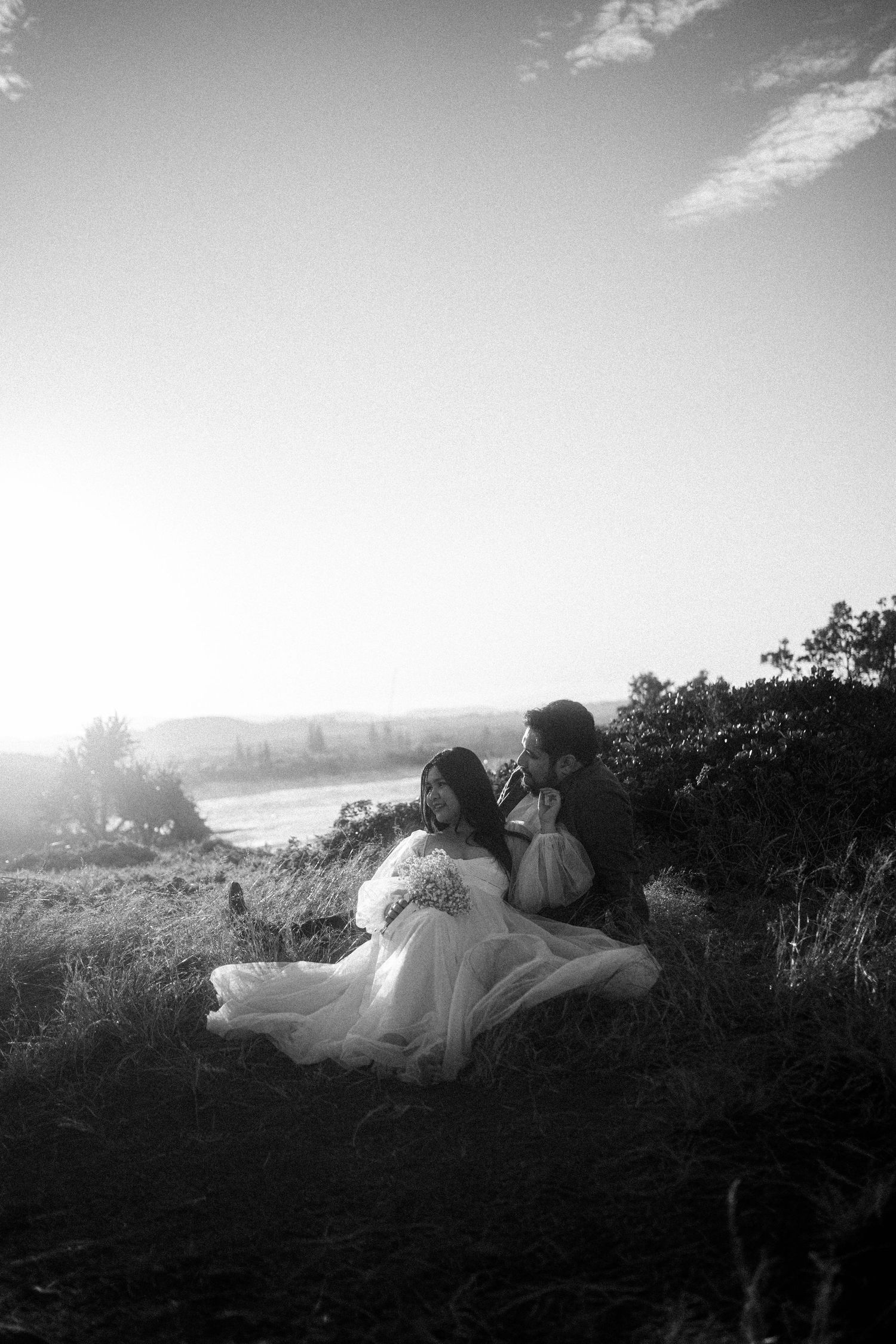 A couple relaxes together on a grassy hillside silhouetted against a moody black and white sky at sunset.