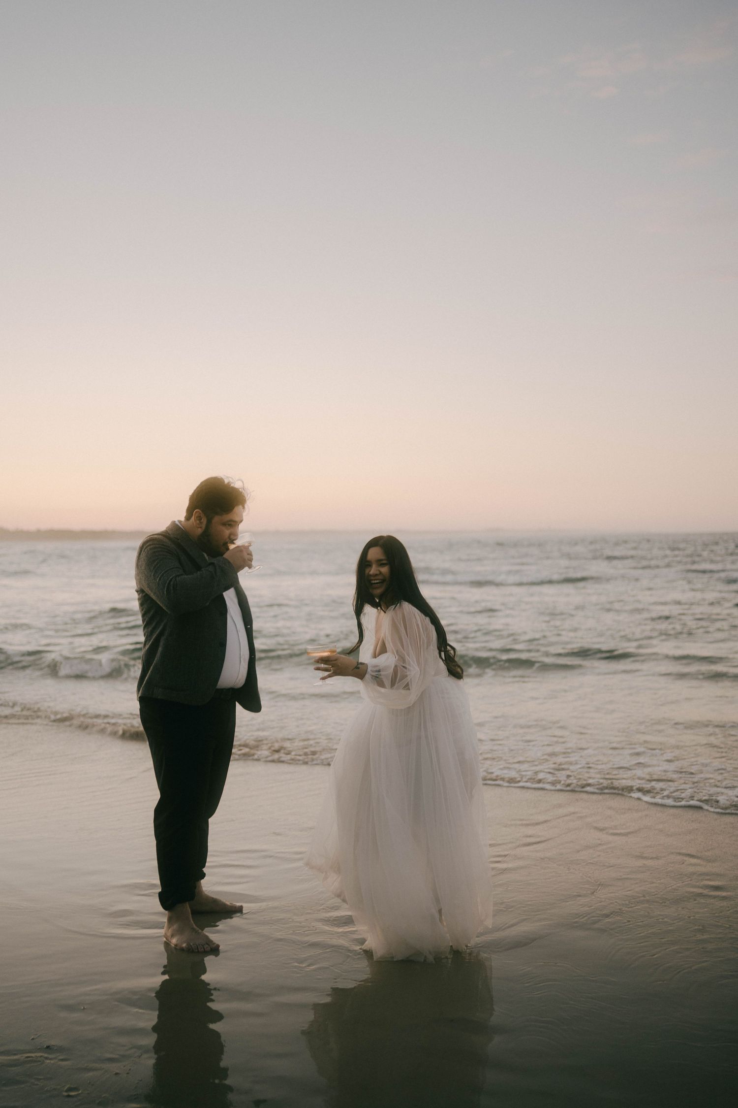 A couple in wedding attire stands at the edge of the ocean during sunset creating a romantic silhouette against the waves.