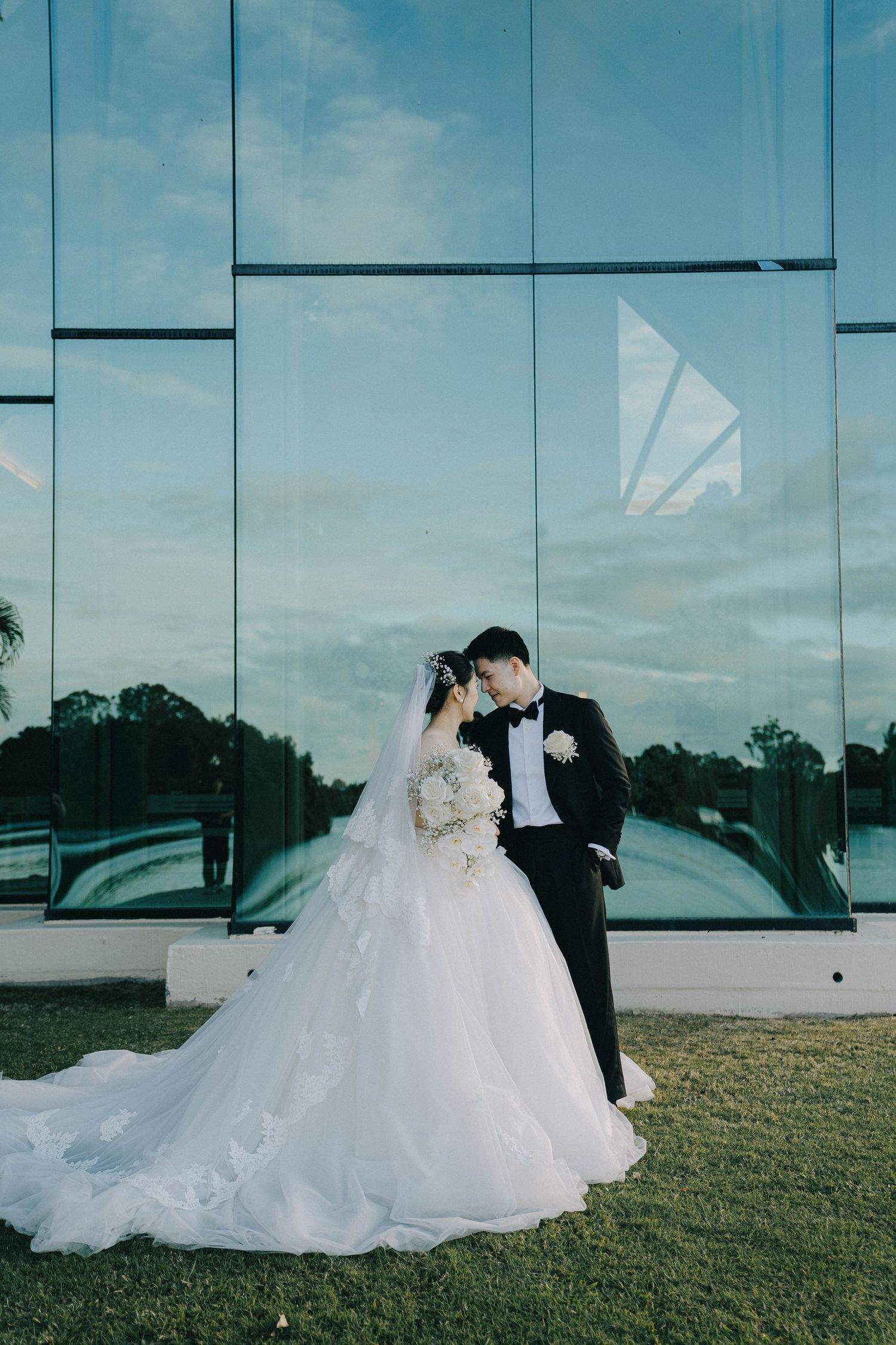 Romantic wedding photos taken on a grassy lawn against a modern glass building with reflective surfaces.
