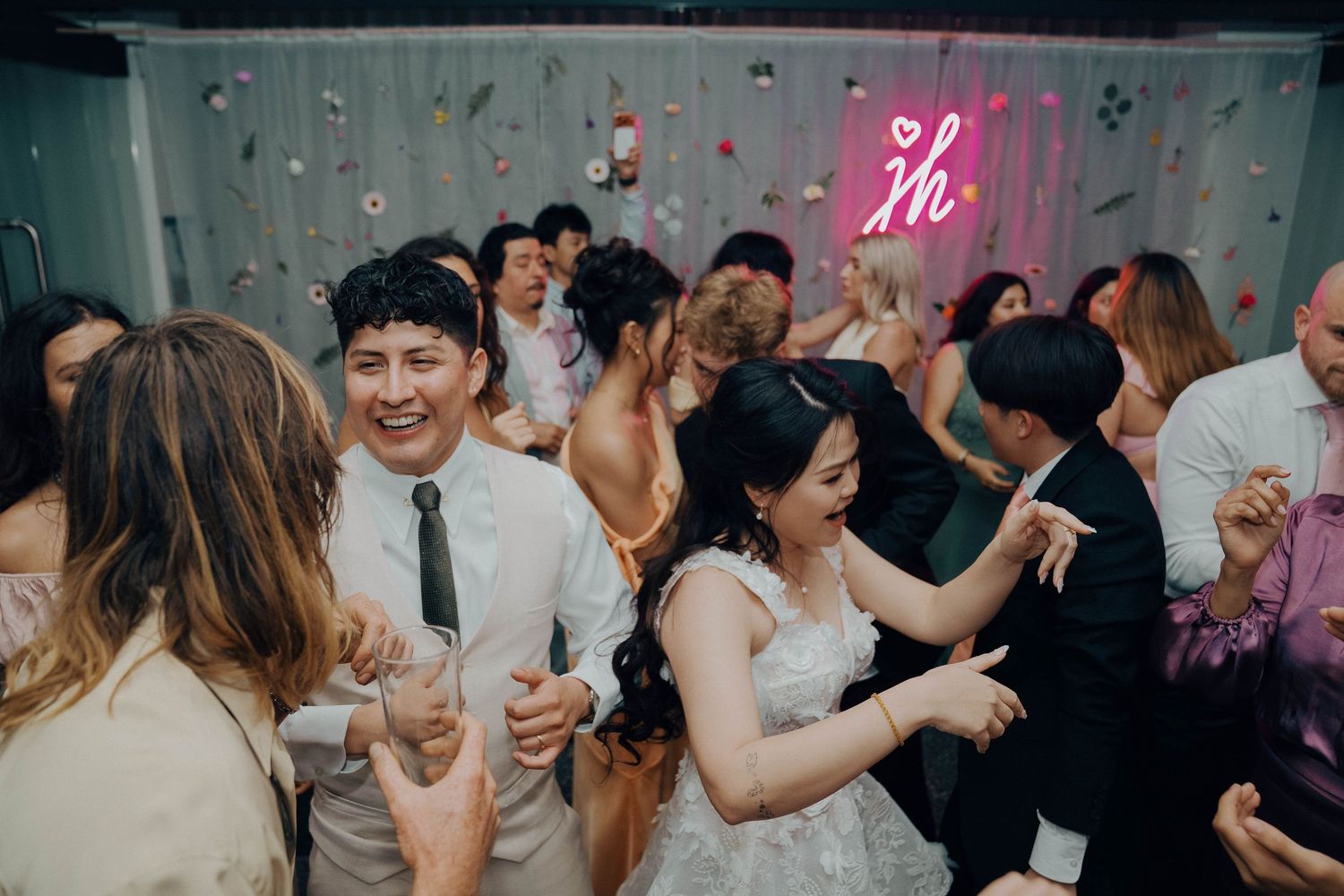 Wedding guests dance and celebrate on a crowded dance floor under pink lighting at an indoor reception party.