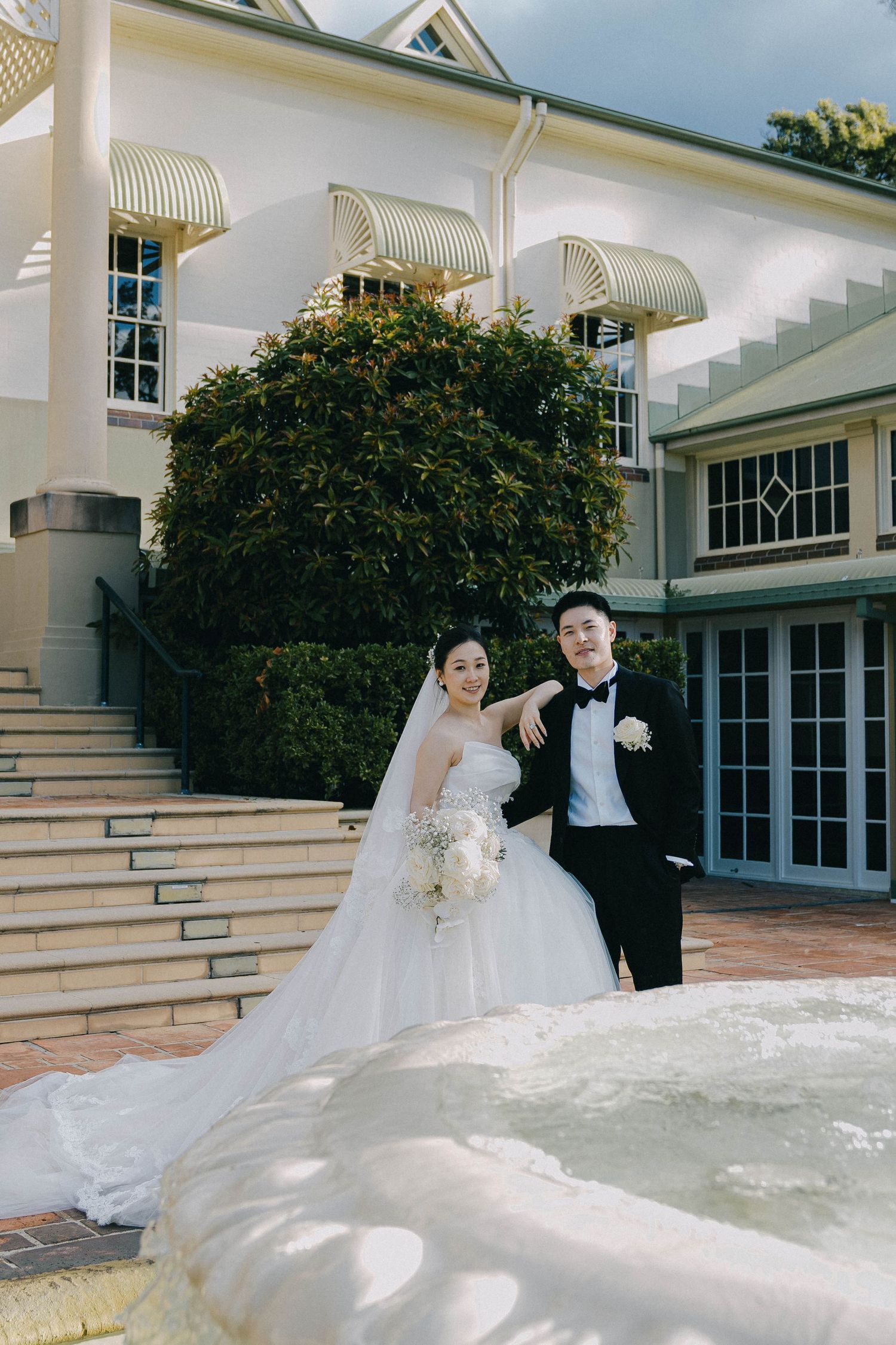 A wedding couple poses on grand white steps outside an elegant building with striped awnings and lush greenery.
