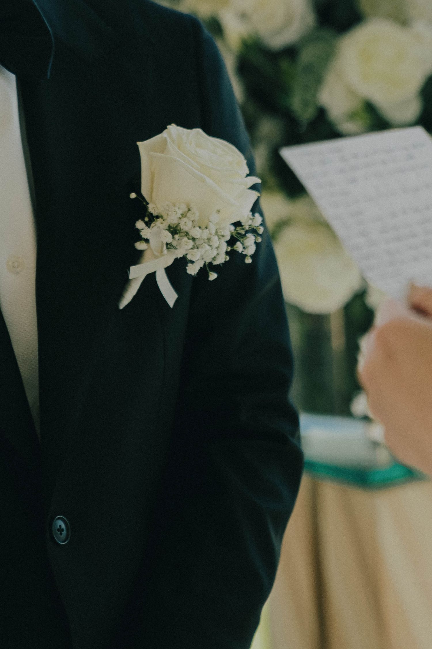 A close-up of white roses and floral decorations with a black suit jacket in the background at a formal event.