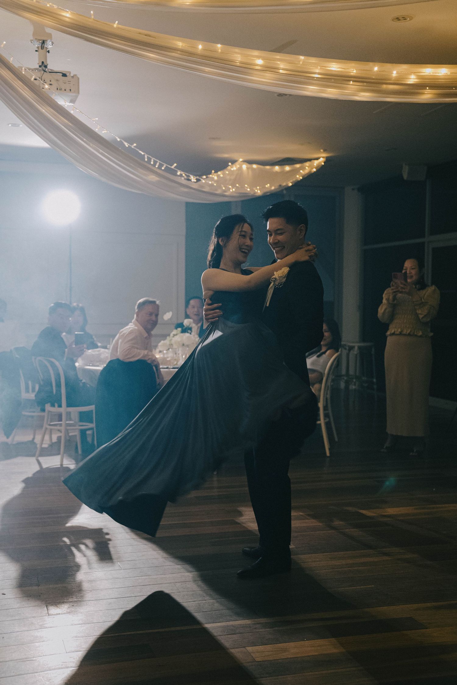 Couples gracefully dance together in a dimly lit ballroom with elegant ceiling lighting and hardwood floors.