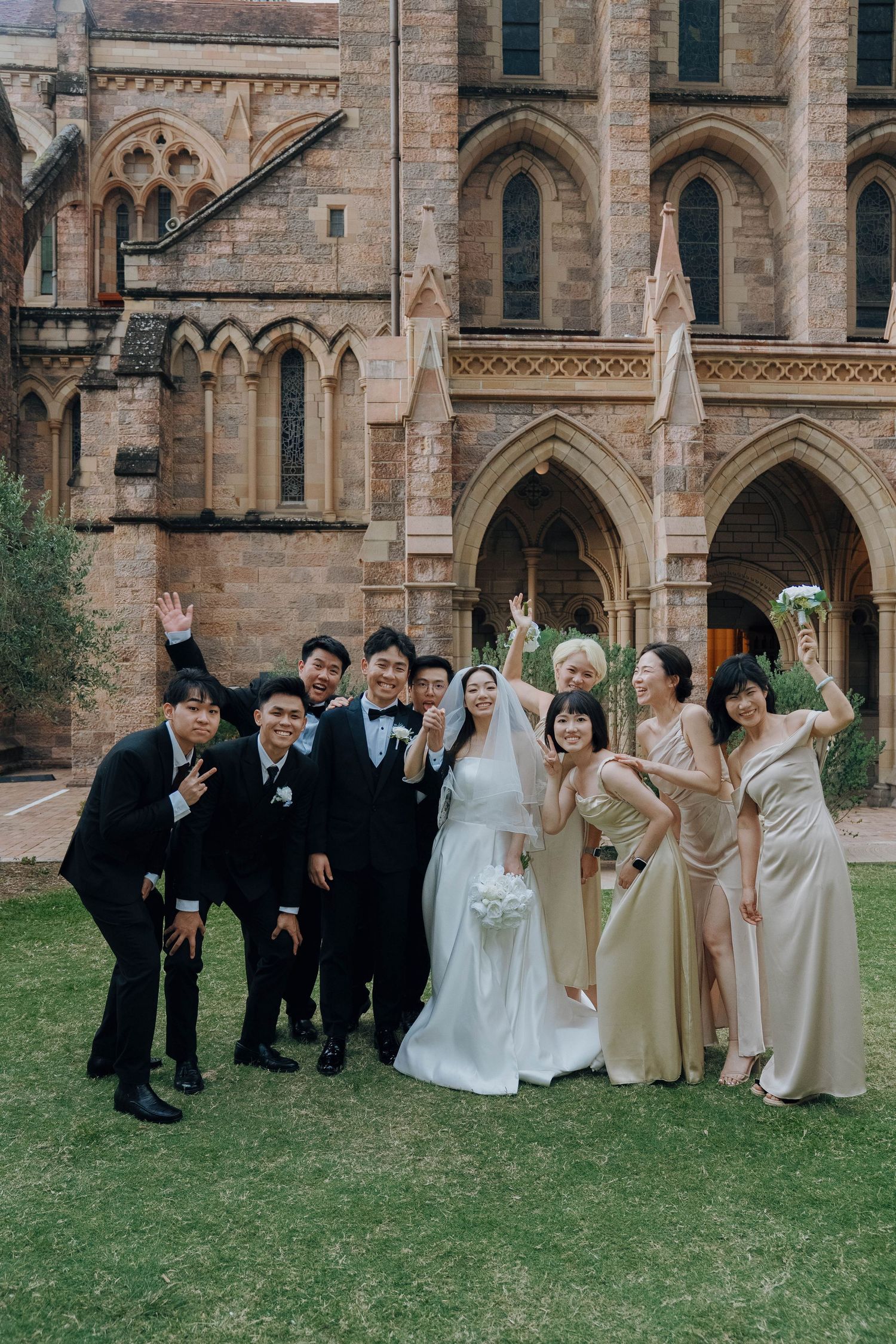 Wedding party poses joyfully on grass in front of gothic stone cathedral with ornate arched architecture.