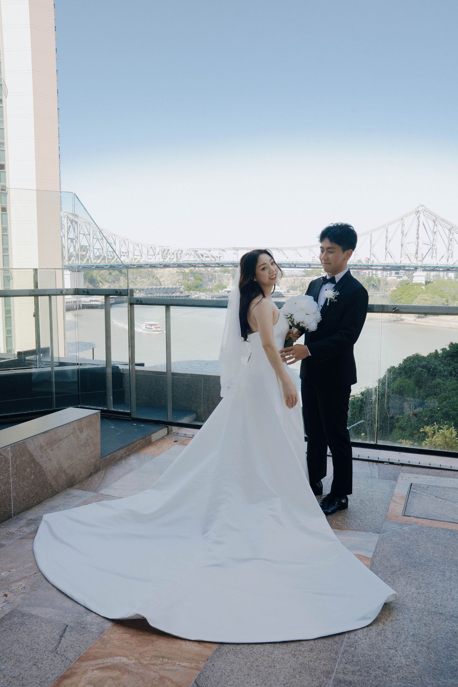 A couple in wedding attire dance on a rooftop terrace with a bridge visible in the background.