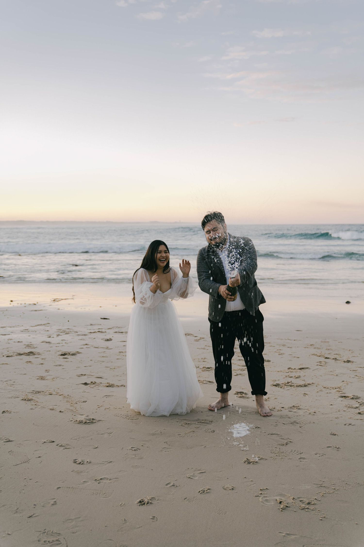 A couple in wedding attire plays together on a sandy beach at sunset, creating joyful moments by the ocean.