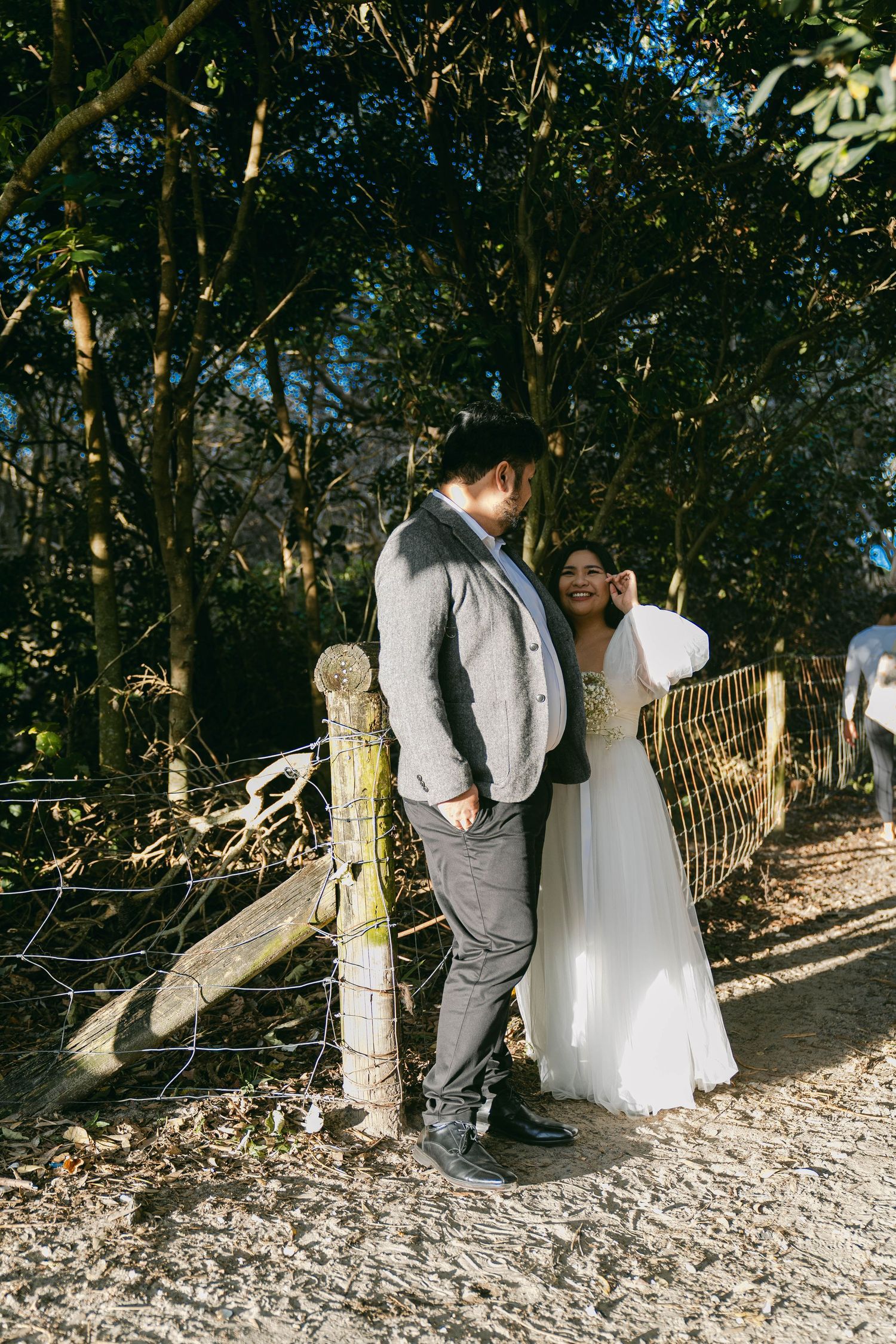 A romantic couple in wedding attire embraces on a rocky beach path surrounded by trees and natural scenery.