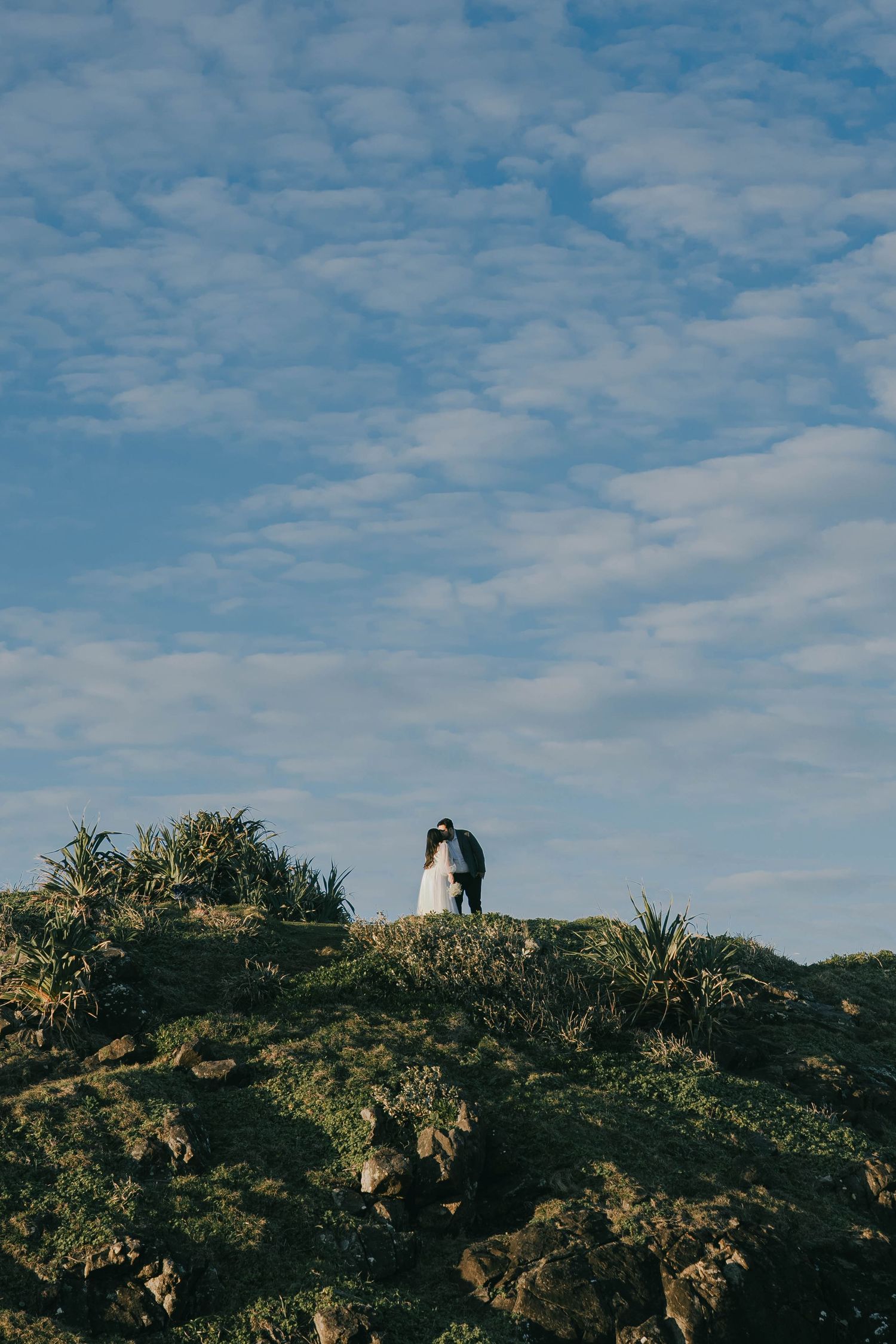 A couple stands together on a hilltop silhouetted against a bright blue sky with scattered white clouds.