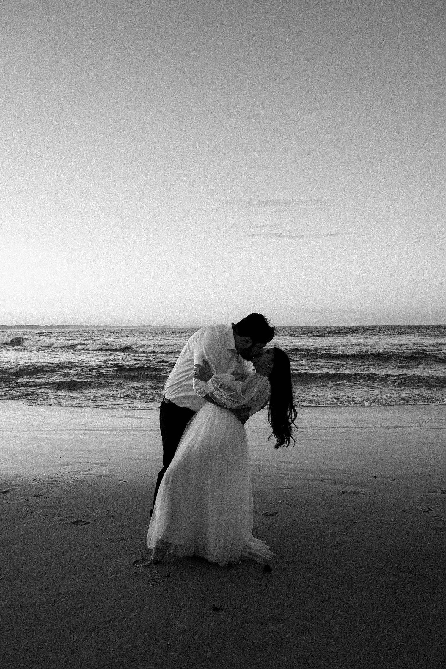A romantic black and white series of photos showing a couple in a white dress embracing on a misty beach shoreline.