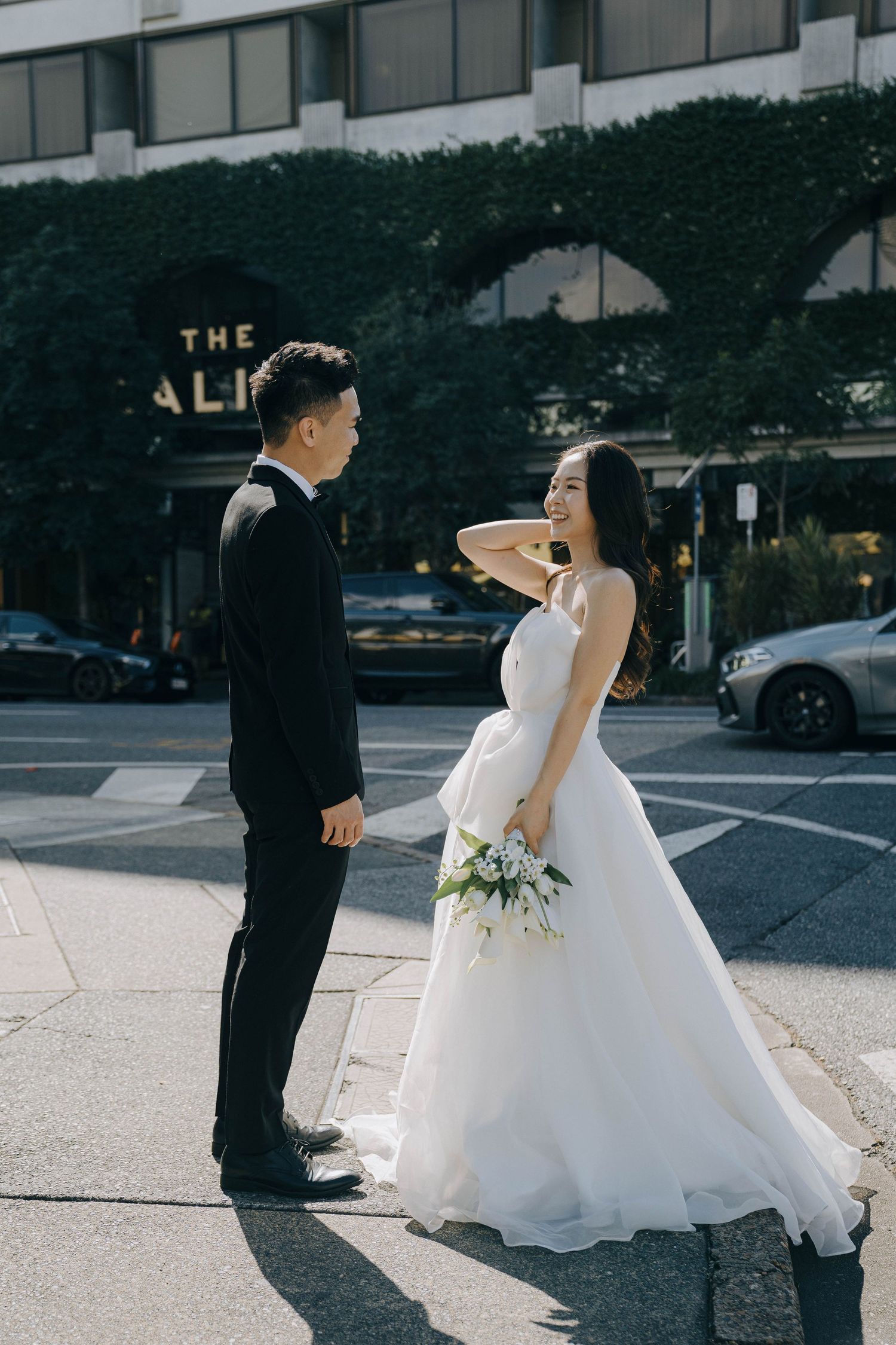 A bride in a flowing white gown and groom in black suit share a romantic first look moment in an urban setting.