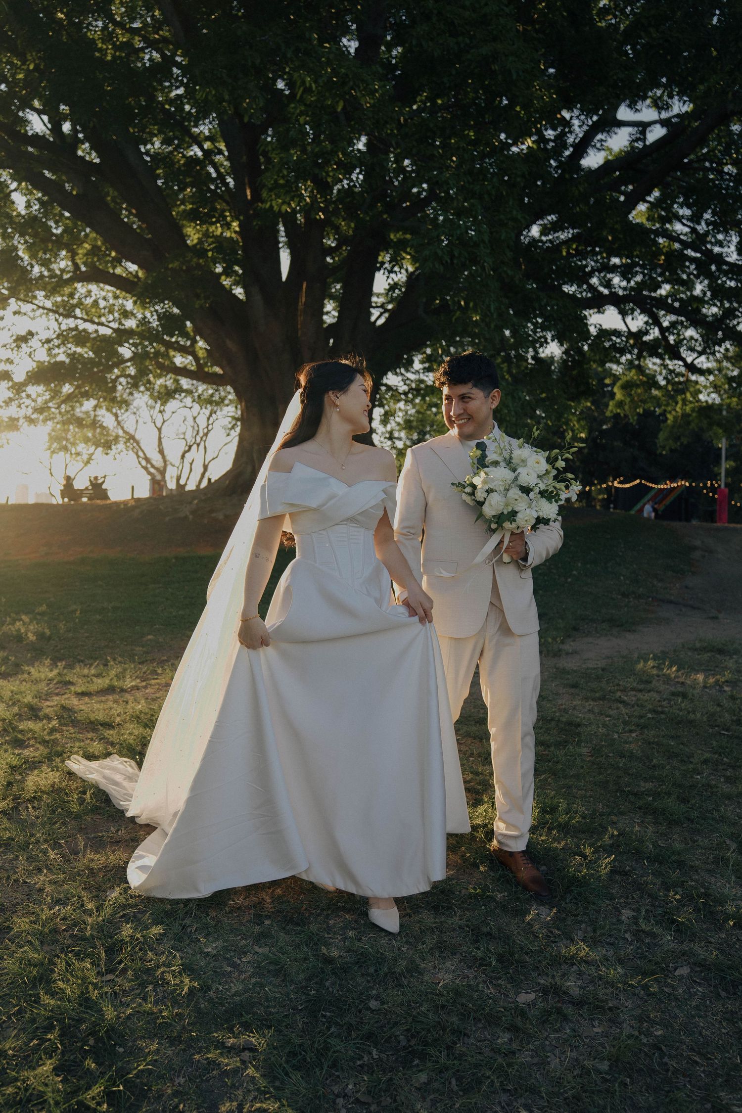 A couple in elegant white attire dance together outdoors at sunset under trees in a romantic setting.