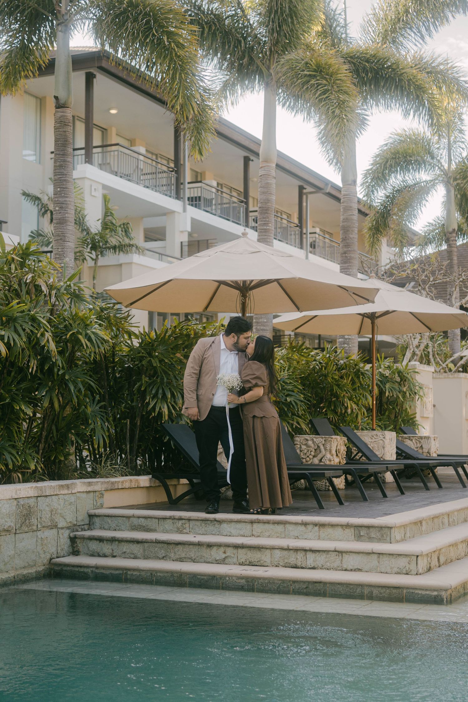 Elegant poolside scene at a luxury resort with palm trees, stone steps, and beige umbrellas beside a tranquil swimming pool.