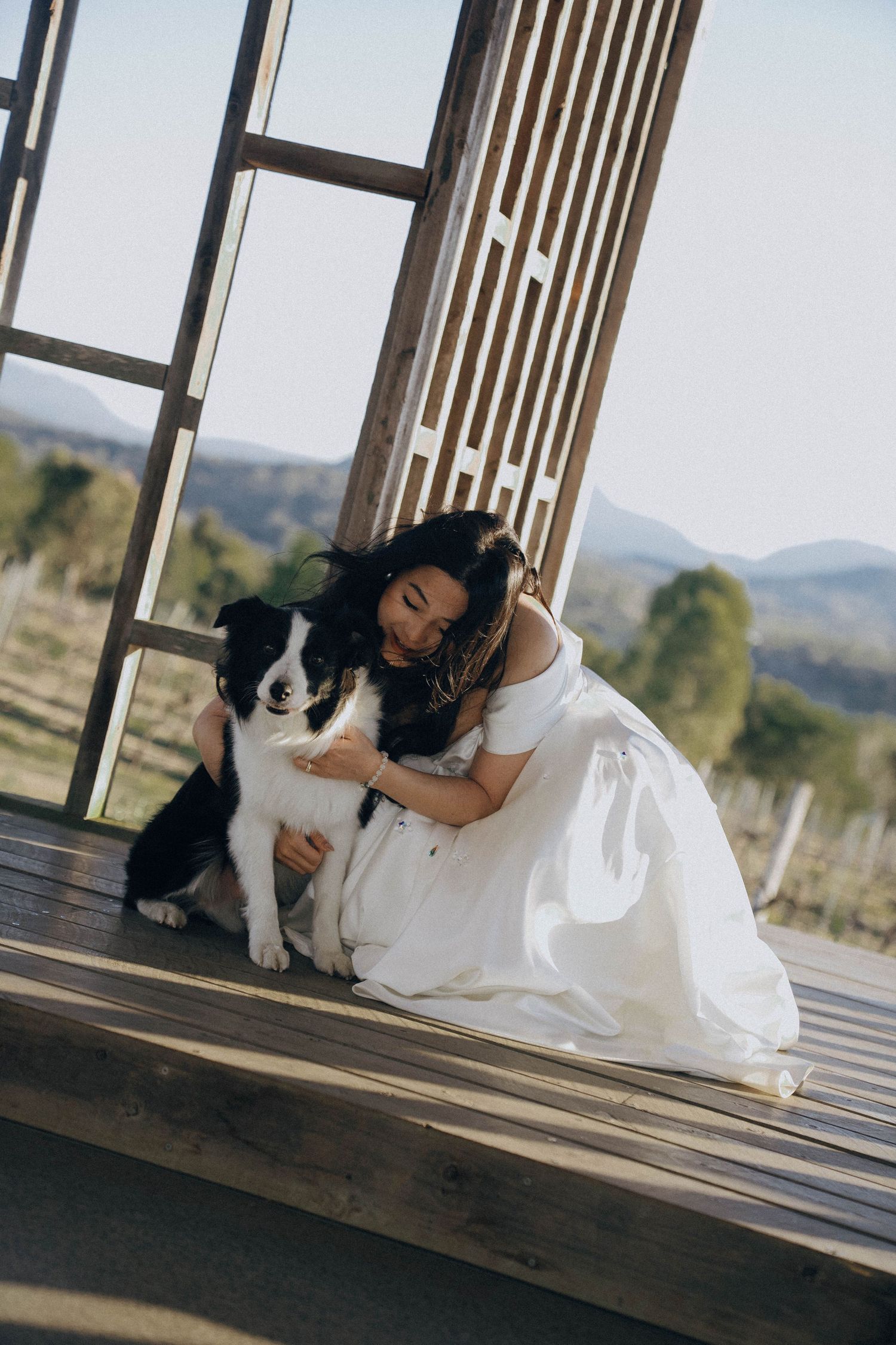 A black and white dog sits with someone wearing a white dress on a wooden deck with mountains in the background.