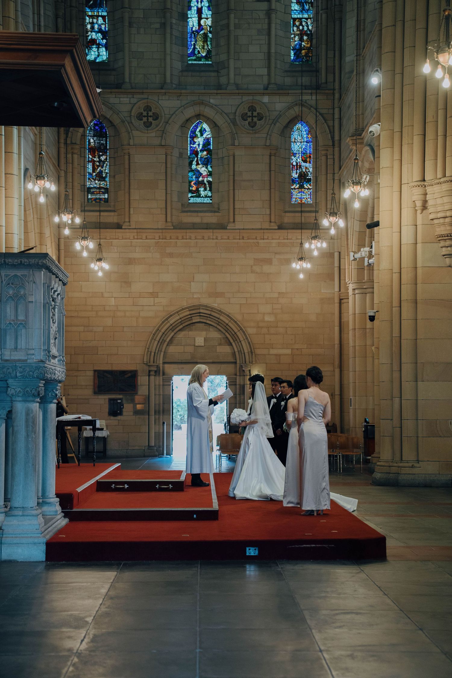 Several people in white robes gather at a cathedral altar with stained glass windows and stone columns in the background.
