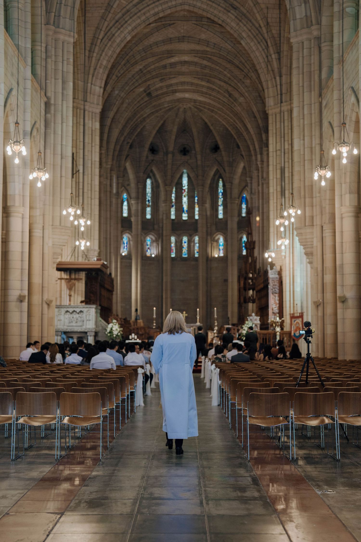 Interior view of a Gothic cathedral with rows of wooden pews and hanging chandeliers illuminating the stone arches.