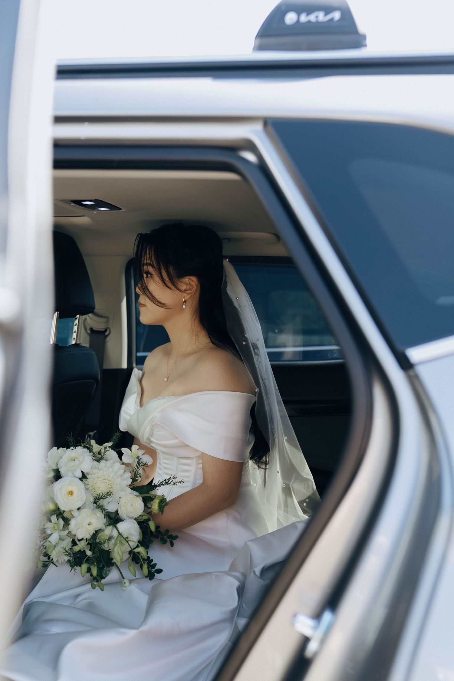 Bride in off-shoulder wedding dress sits elegantly in white car holding a white and green floral bouquet.