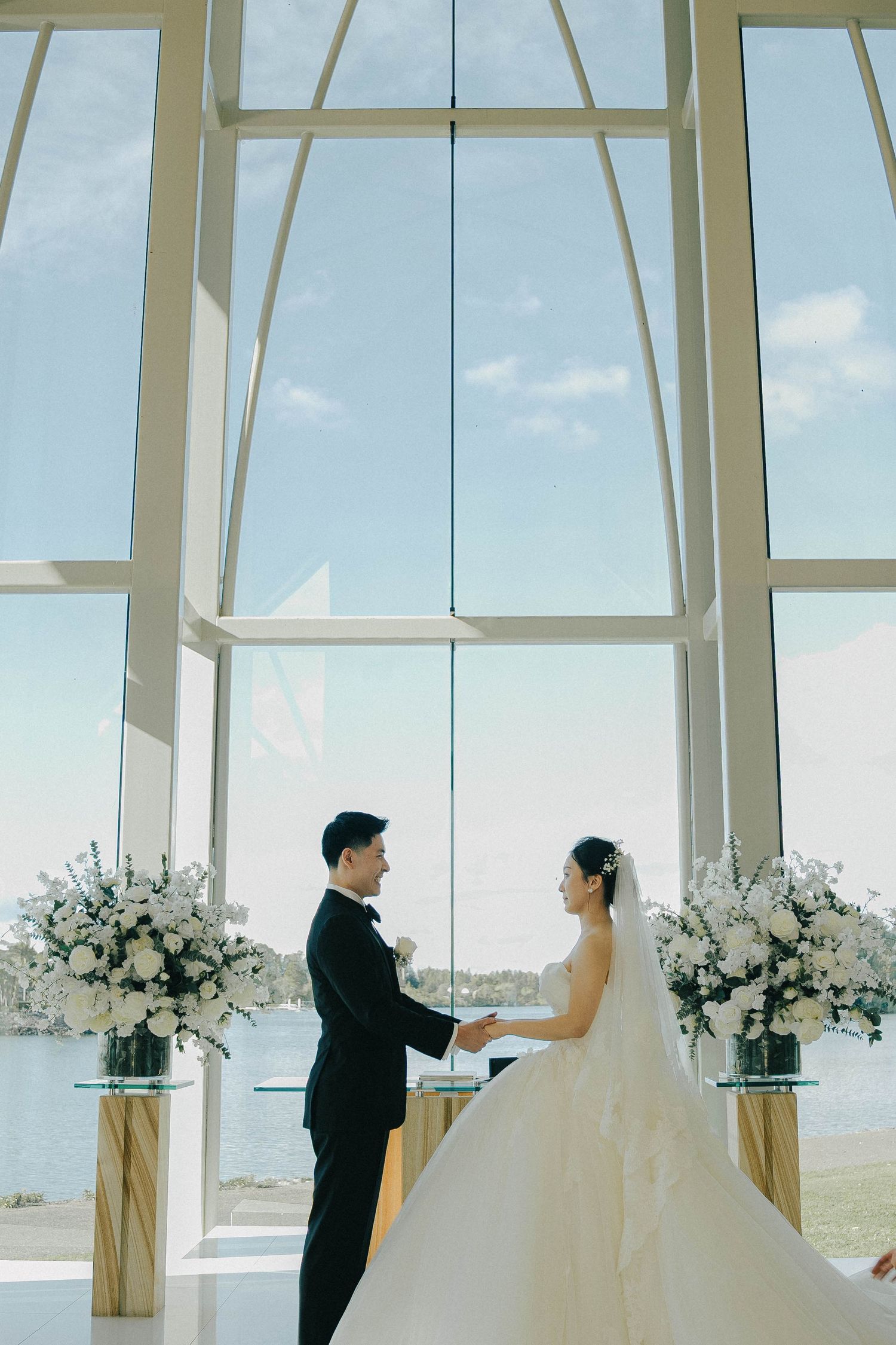 A wedding couple shares a romantic moment in a bright, modern chapel with floor-to-ceiling windows and white floral arrangements.