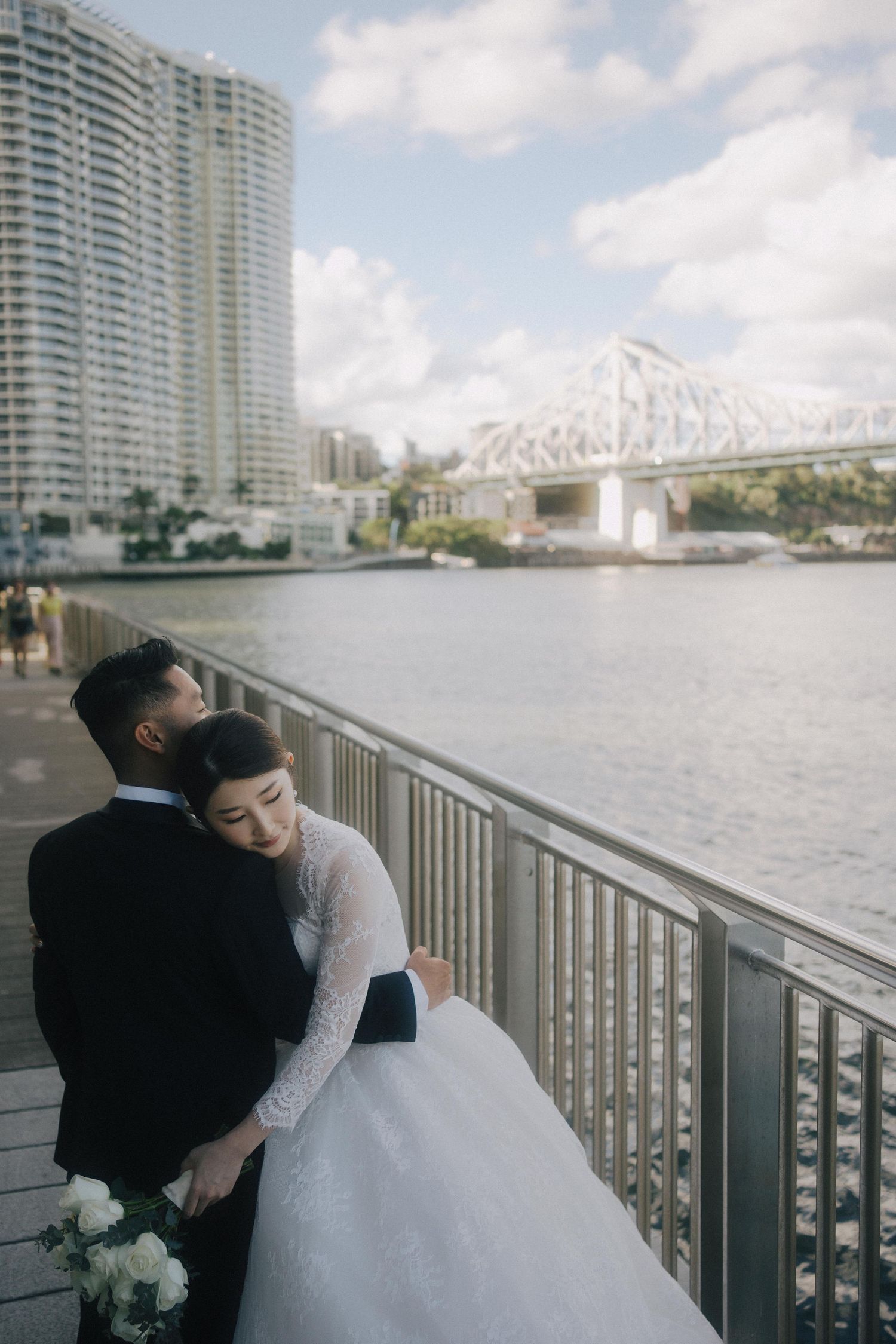 A couple in wedding attire embraces on a riverside promenade with the Story Bridge and high-rise buildings in Brisbane.