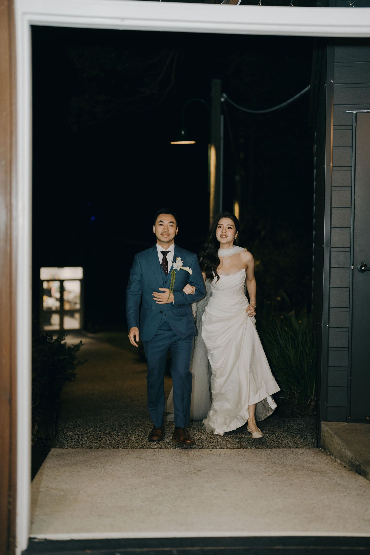 A couple walks together in wedding attire, the bride in white and groom in navy suit, through a dimly lit venue entrance.