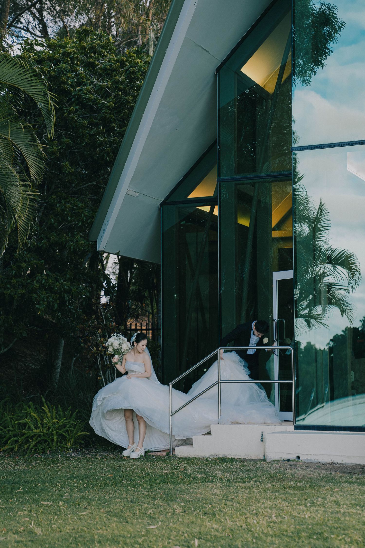 A flowing white wedding dress billows in the breeze outside a modern glass chapel with tropical greenery surroundings.