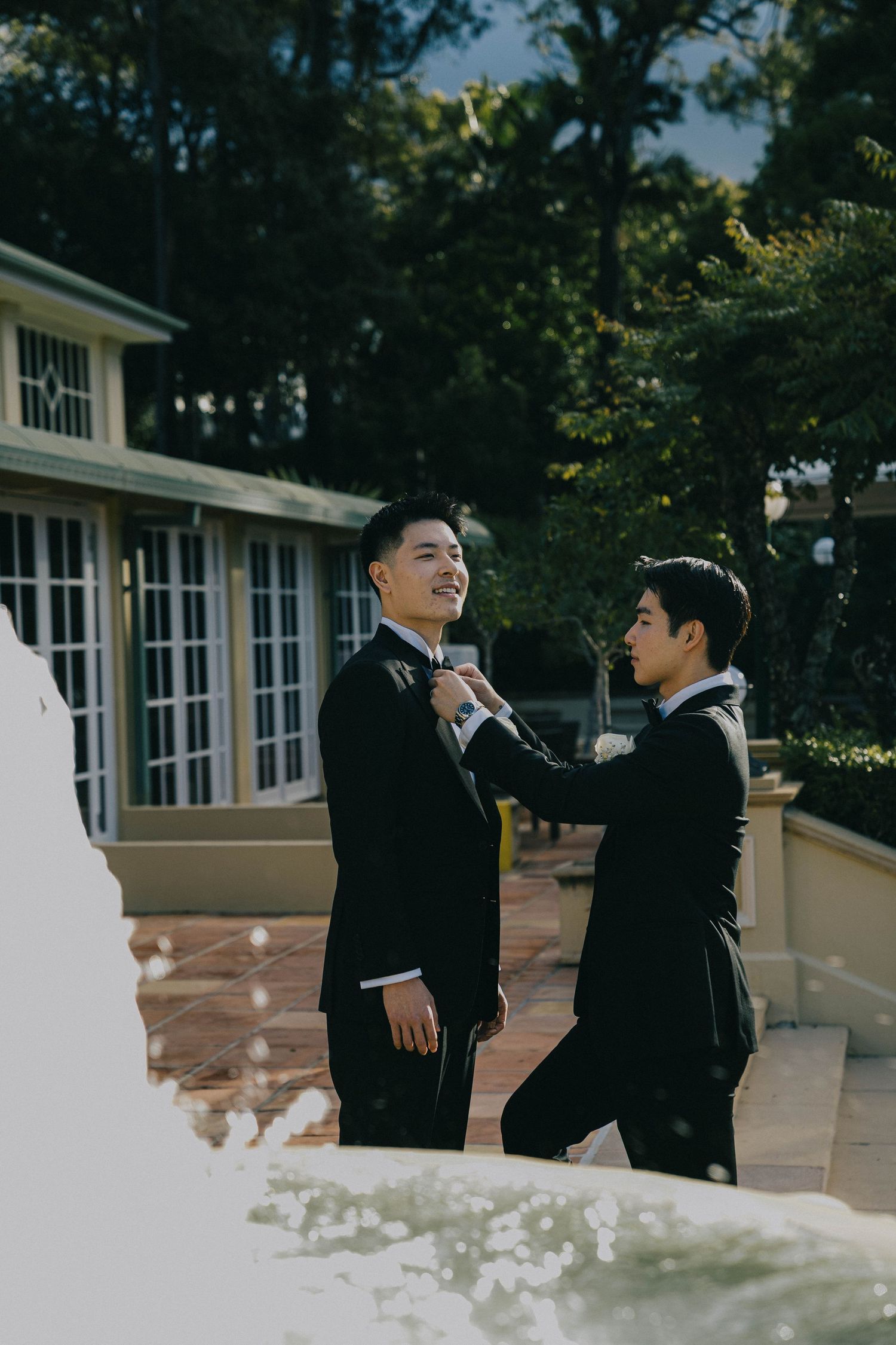 Two people in black suits playfully interact outdoors near a white building with windows in a candid photo series.