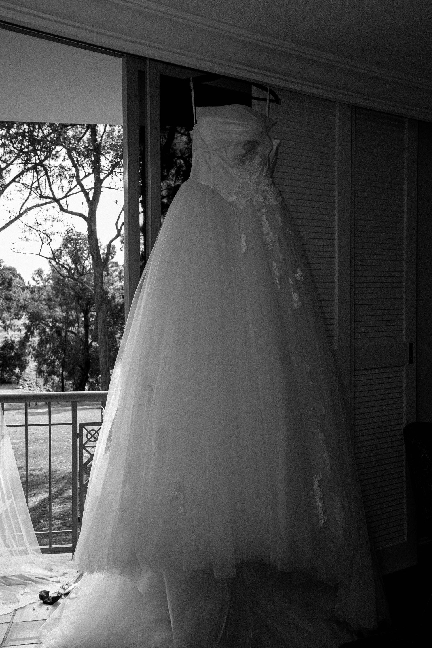 A white wedding dress with tulle skirt and beaded bodice hangs elegantly by a window in black and white photography.