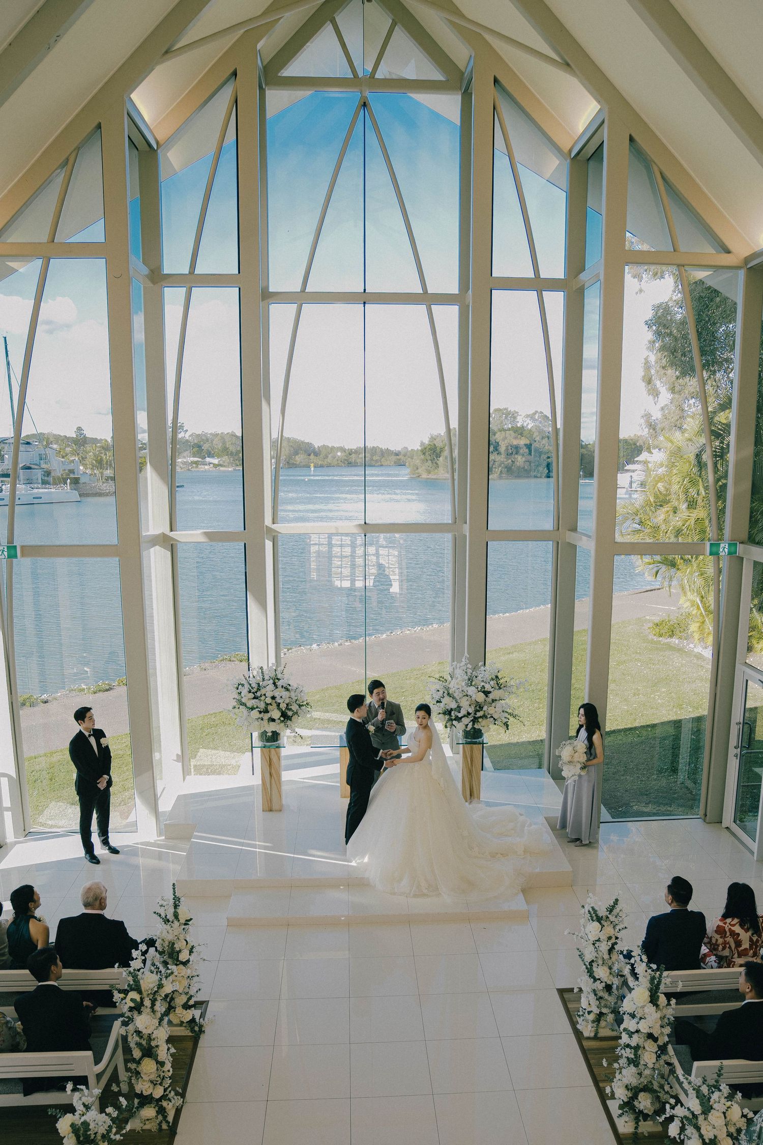 A wedding ceremony in a stunning chapel with floor-to-ceiling windows overlooking a serene lake and lush landscape.