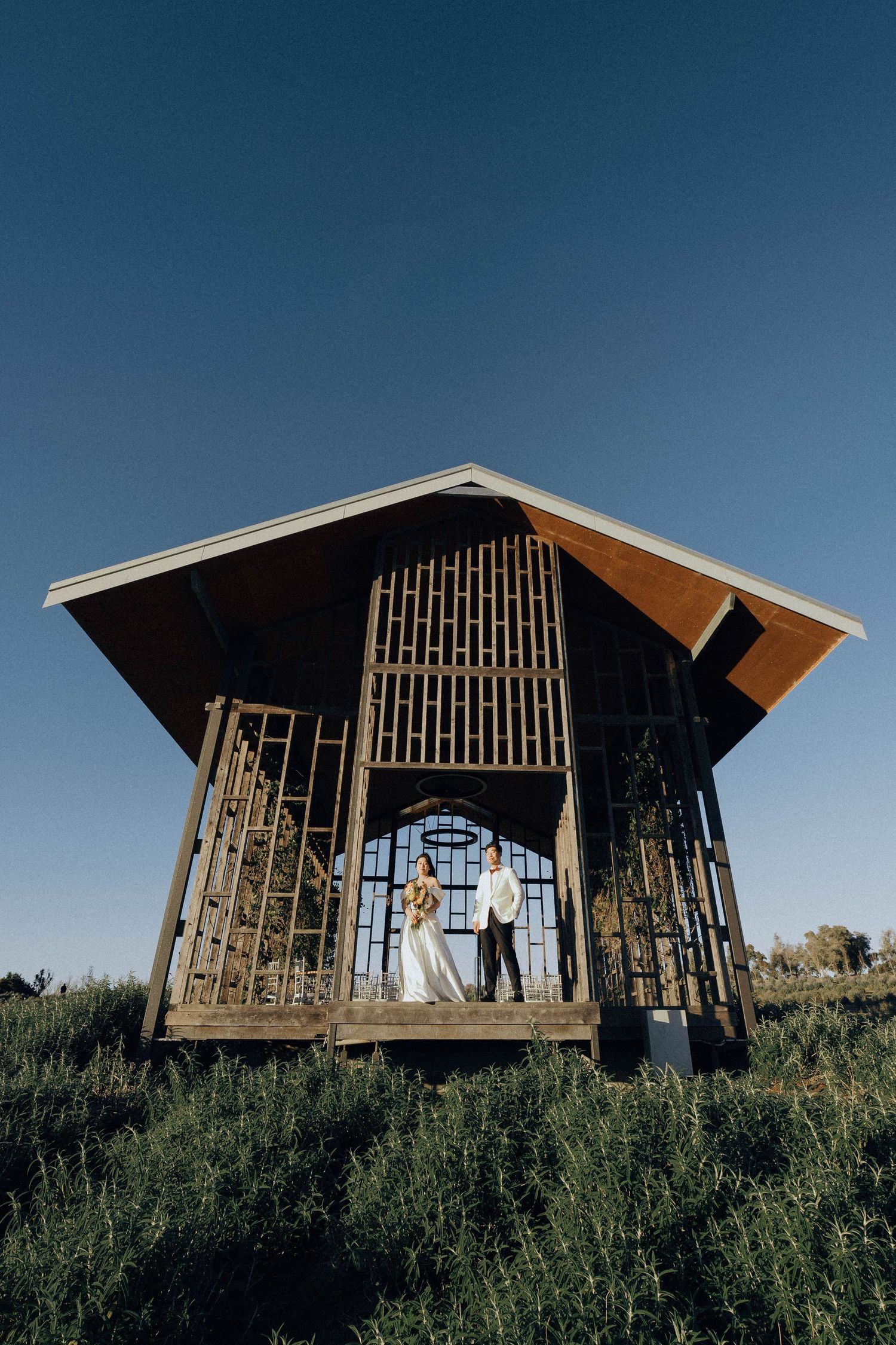 A modern wooden pavilion structure with slatted walls stands against a blue sky on a grassy hillside.