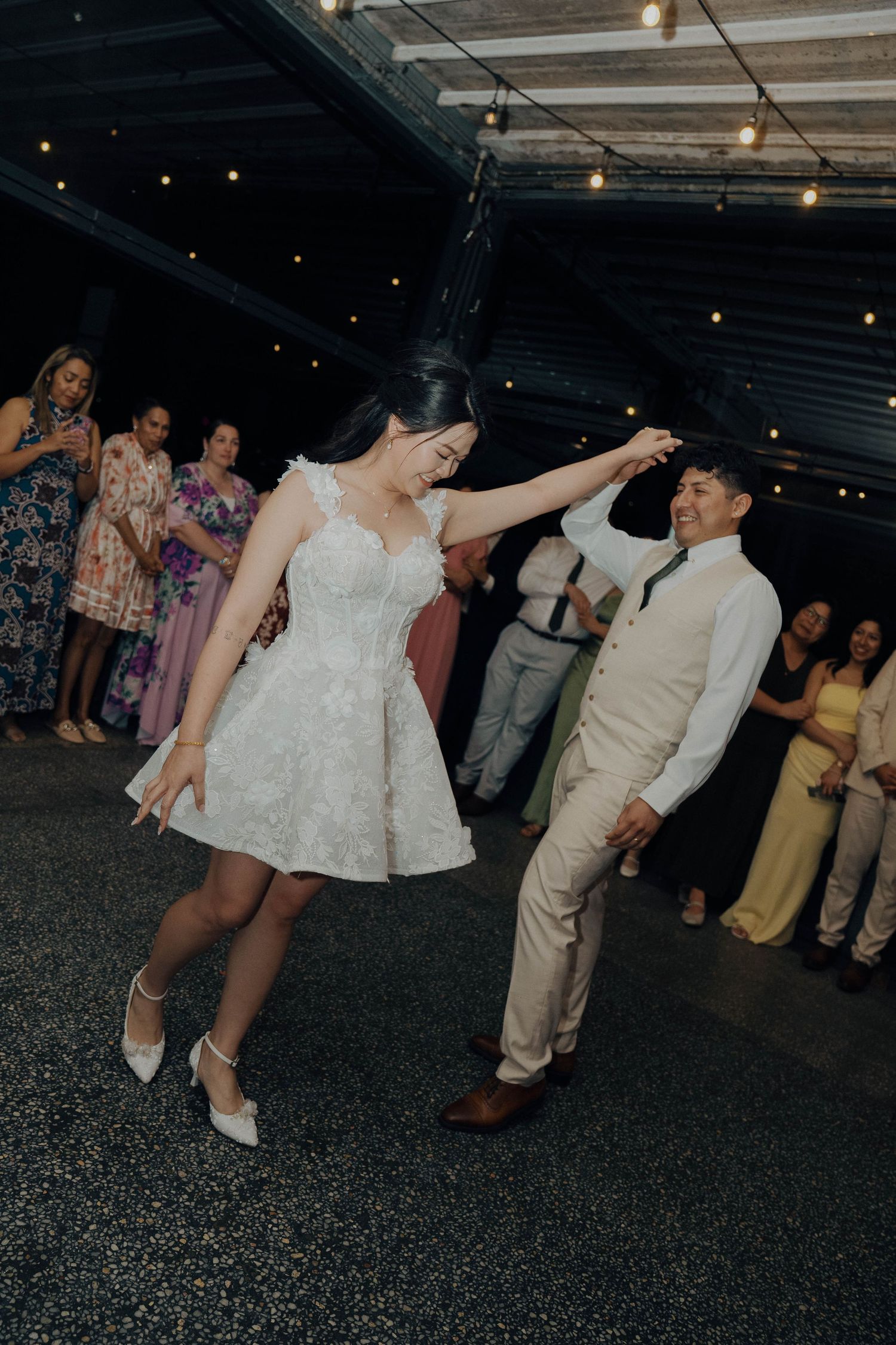 Wedding guests dance and celebrate on a dark dance floor under string lights at an evening reception.