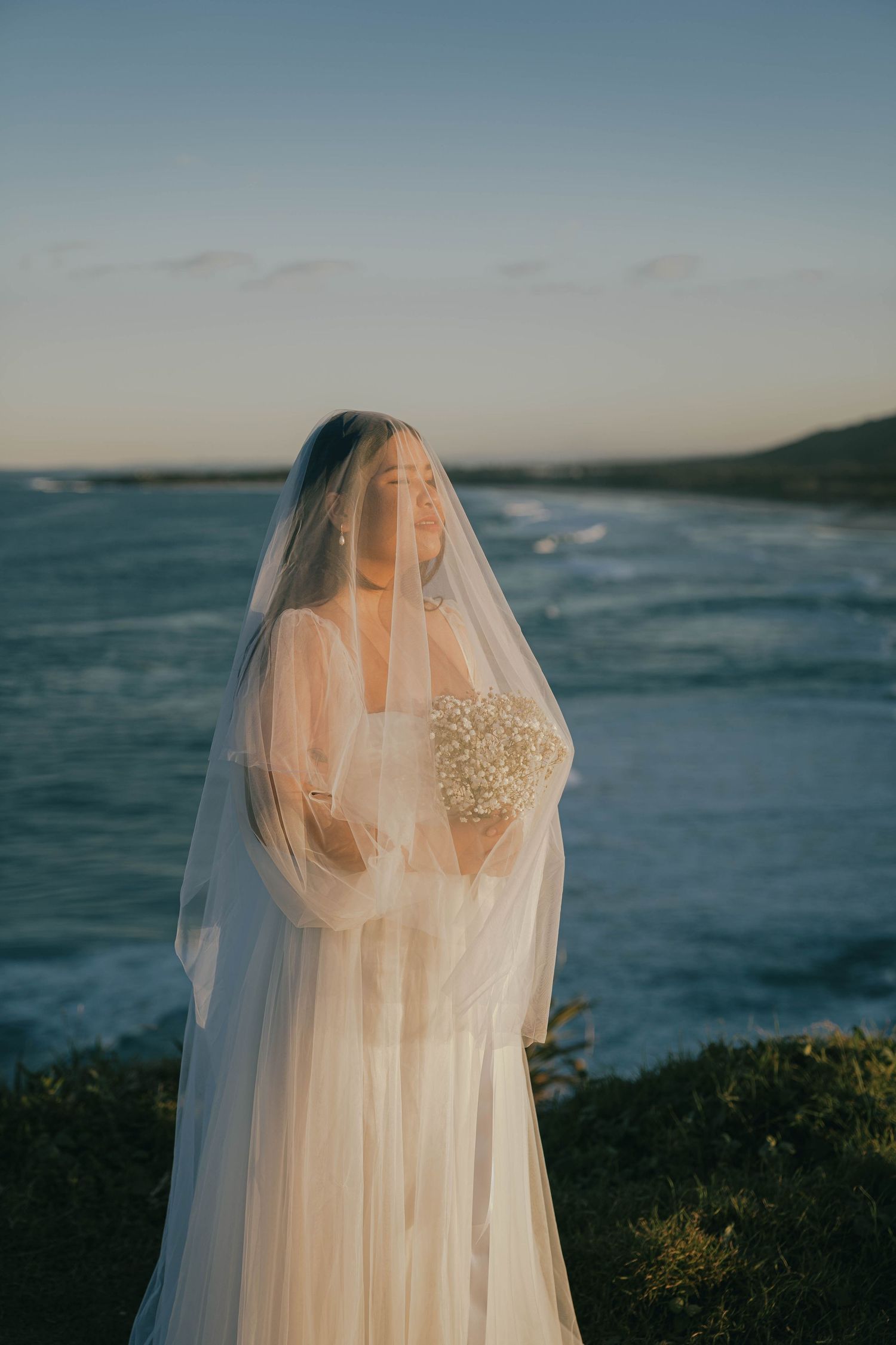 A bride in a flowing white veil stands against a scenic ocean backdrop at sunset.