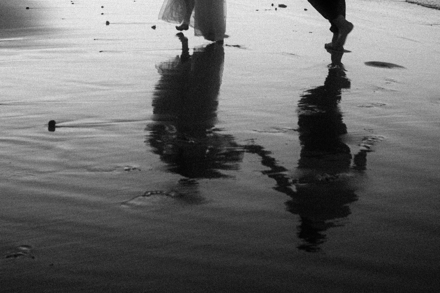 Silhouettes of people walking on a reflective beach surface at sunset with rippling water in black and white.