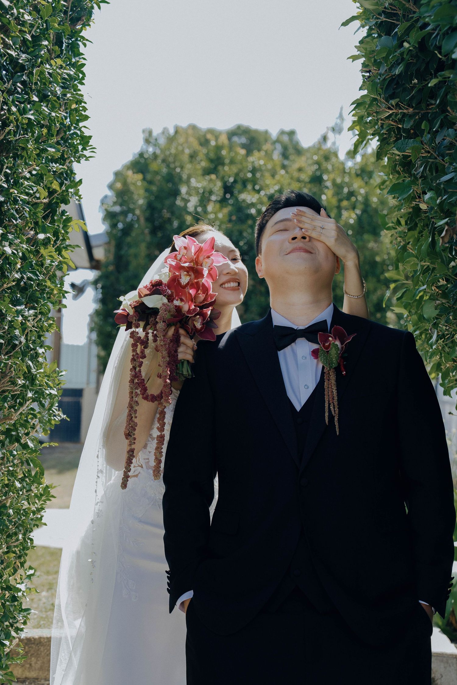 A couple at their wedding ceremony with a pink bouquet and formal attire standing between green hedges outdoors.