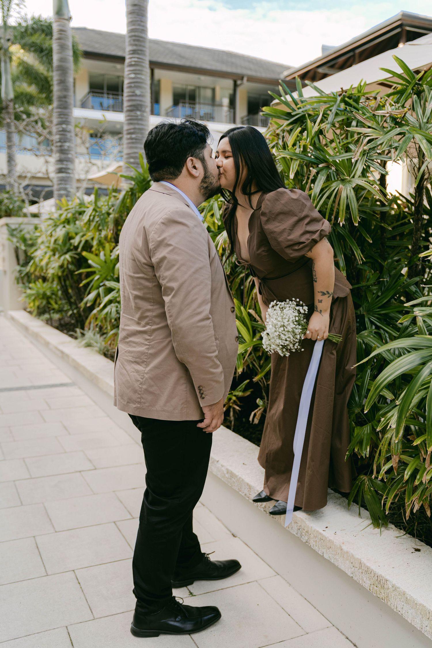 A romantic couple sharing a tender moment together against a tropical garden backdrop with palm trees and white walls.